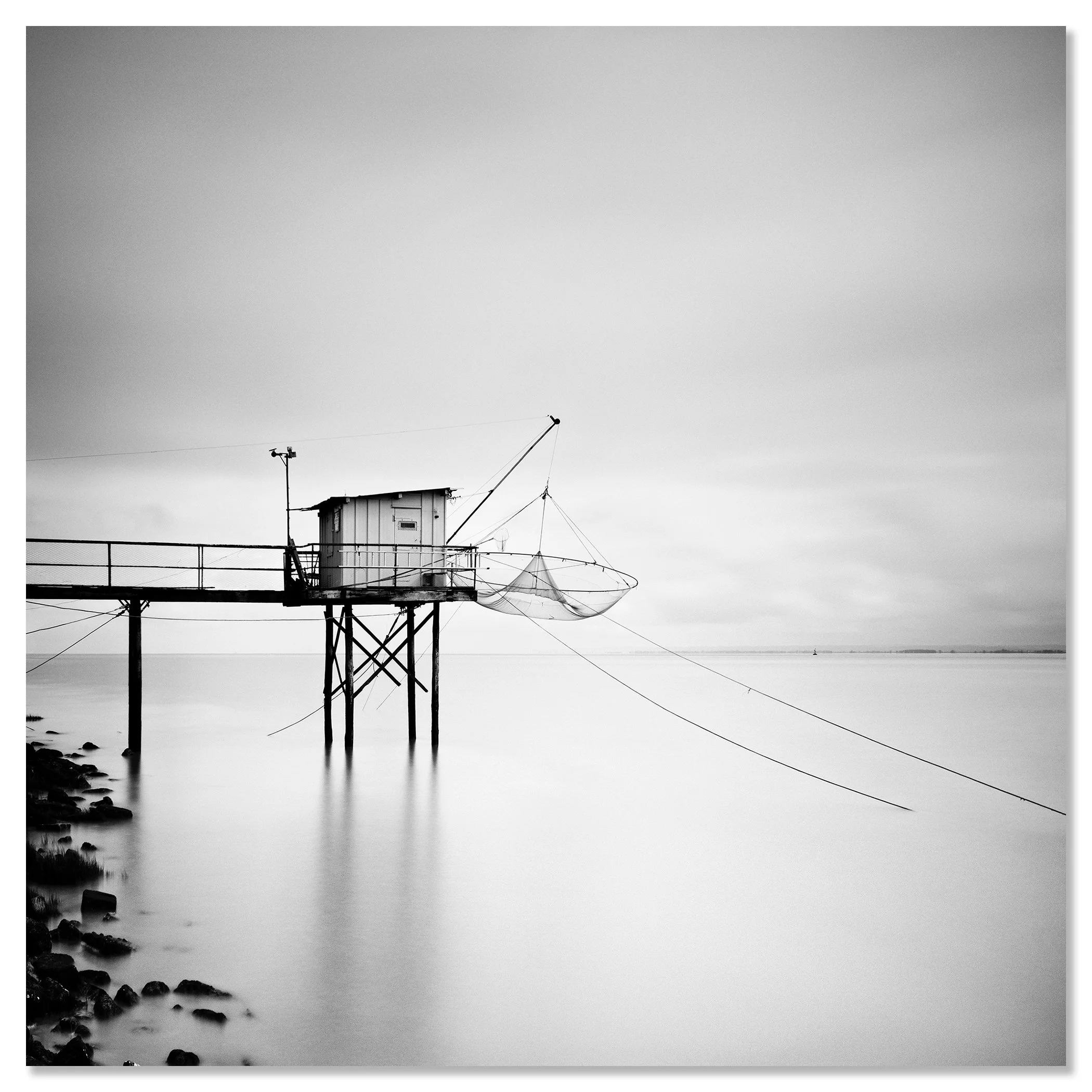 Black-and-white photo of a stilted fishing hut with a net over calm water beside a rocky shoreline – dibond frameless