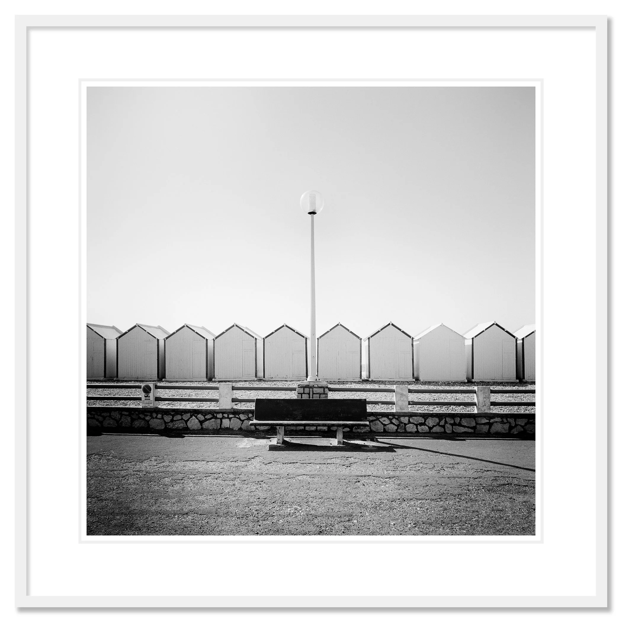 Empty bench on the promenade facing beach huts, centred streetlamp, minimalist black-and-white coastal photograph, Classic frame white