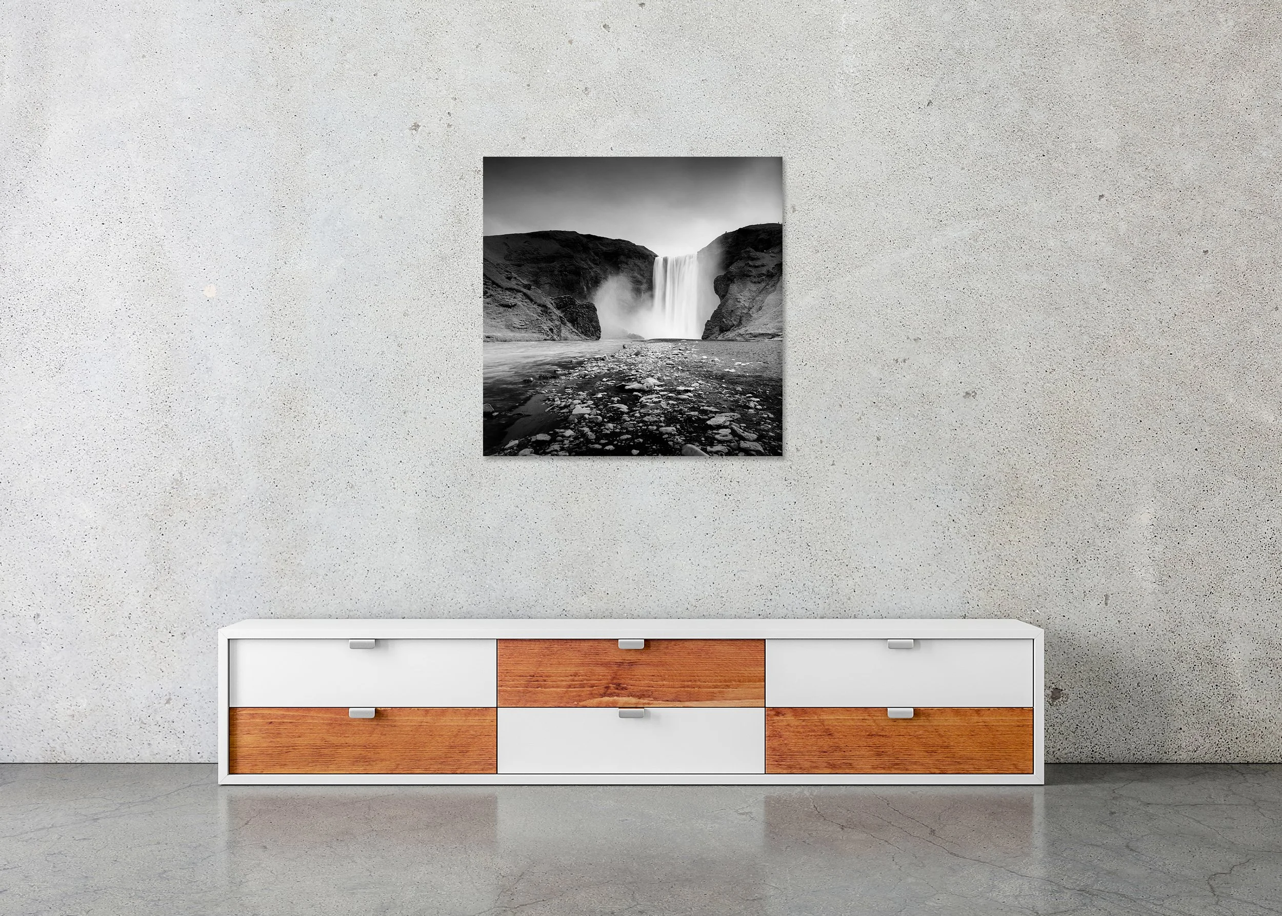 Black and white photograph of Skogafoss waterfall in Iceland, cascading between dark volcanic cliffs with a rocky riverbed in the foreground; frameless photo print mounted to Dibond.