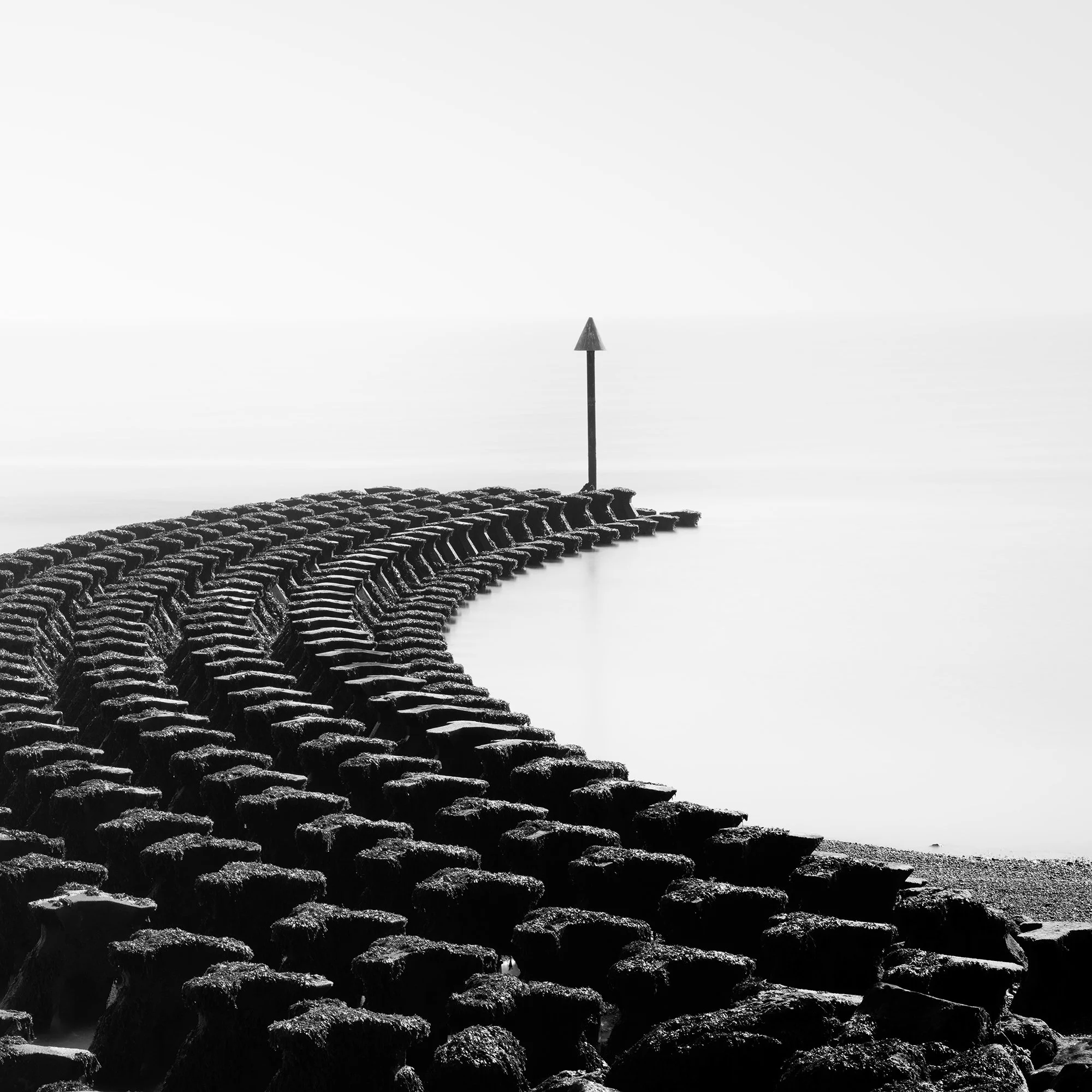 Gerald Berghammer - Black and white minimalist photography. A curved stone jetty extending into a calm body of water, with a tall navigation marker at the end. Print detail 2