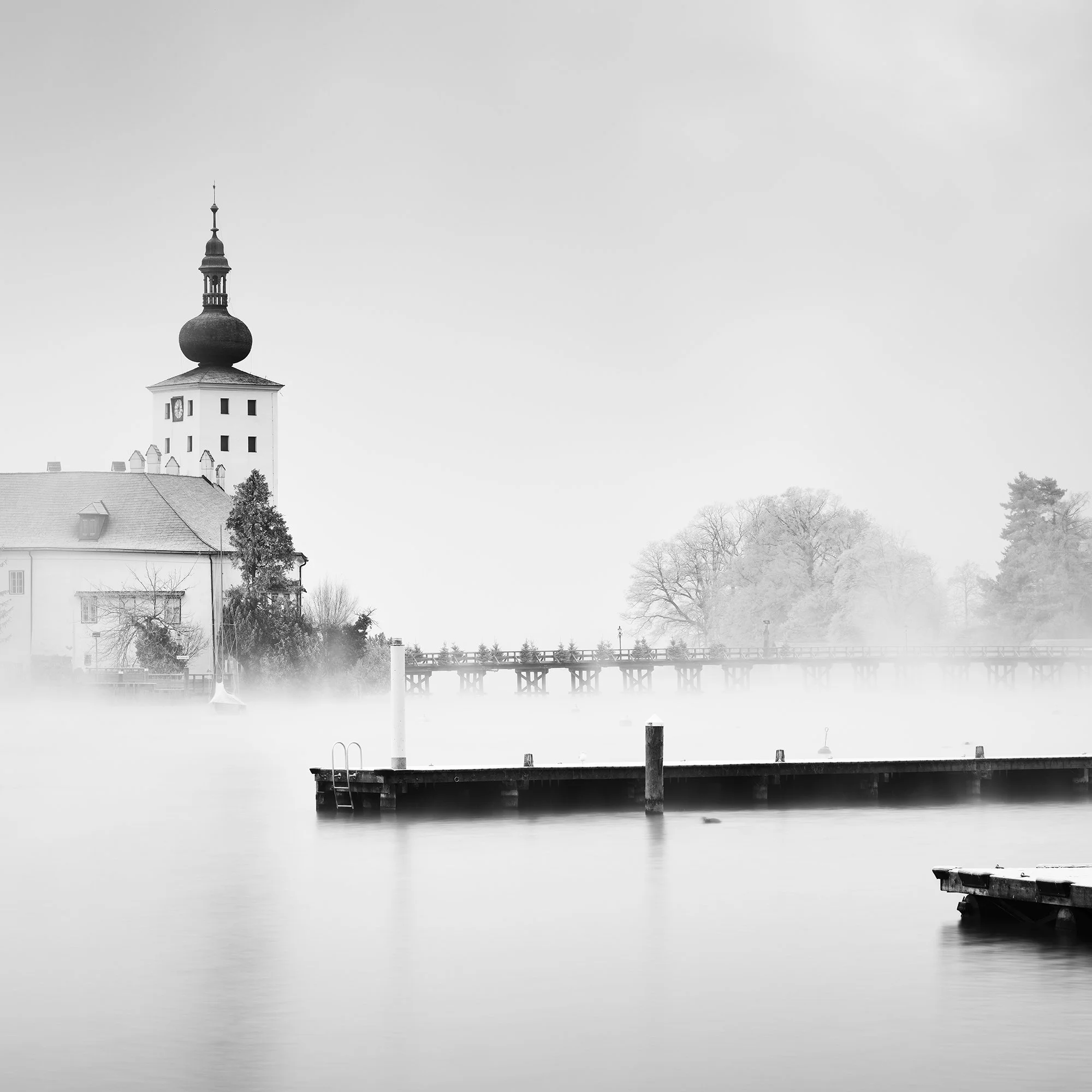 © 2022 Gerald Berghammer - Black and white cityscape photography. Schloss Ort - The historic church surrounded by mist over a lake with docks in the foreground. Print detail 2