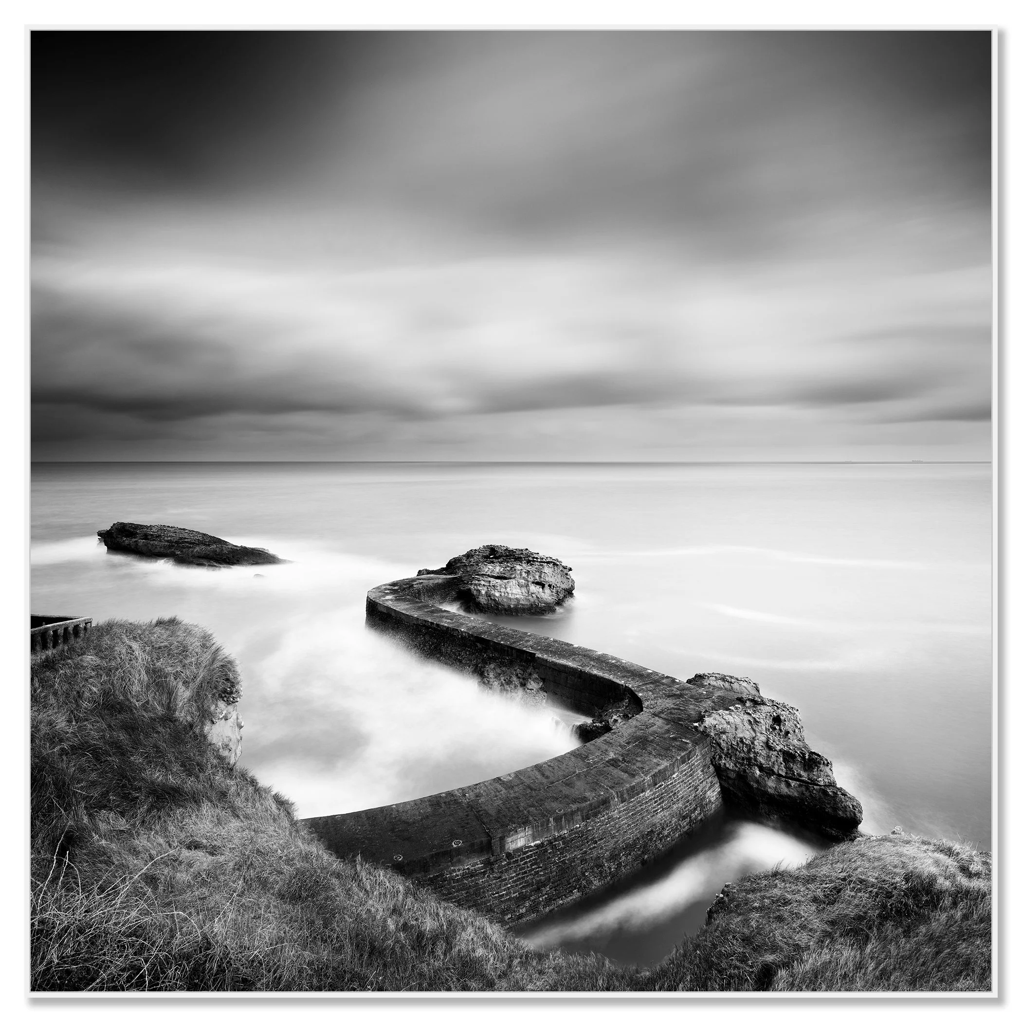 Black and white long-exposure seascape with a curved breakwater pier and rocks on a cloudy coast – framed ArtBox white