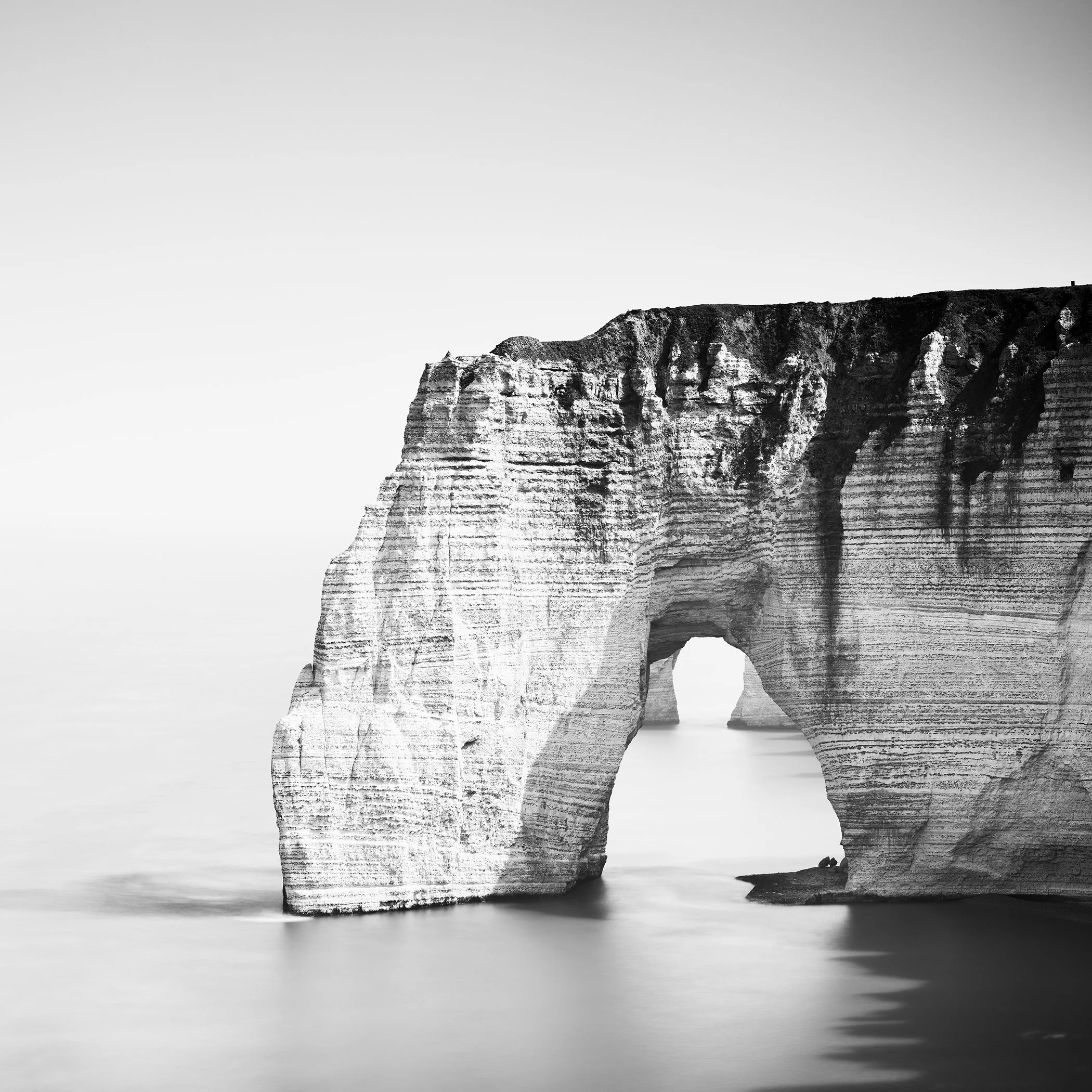 Minimalist monochrome seascape of the Étretat sea arch, layered cliffs and calm water by Gerald Berghammer