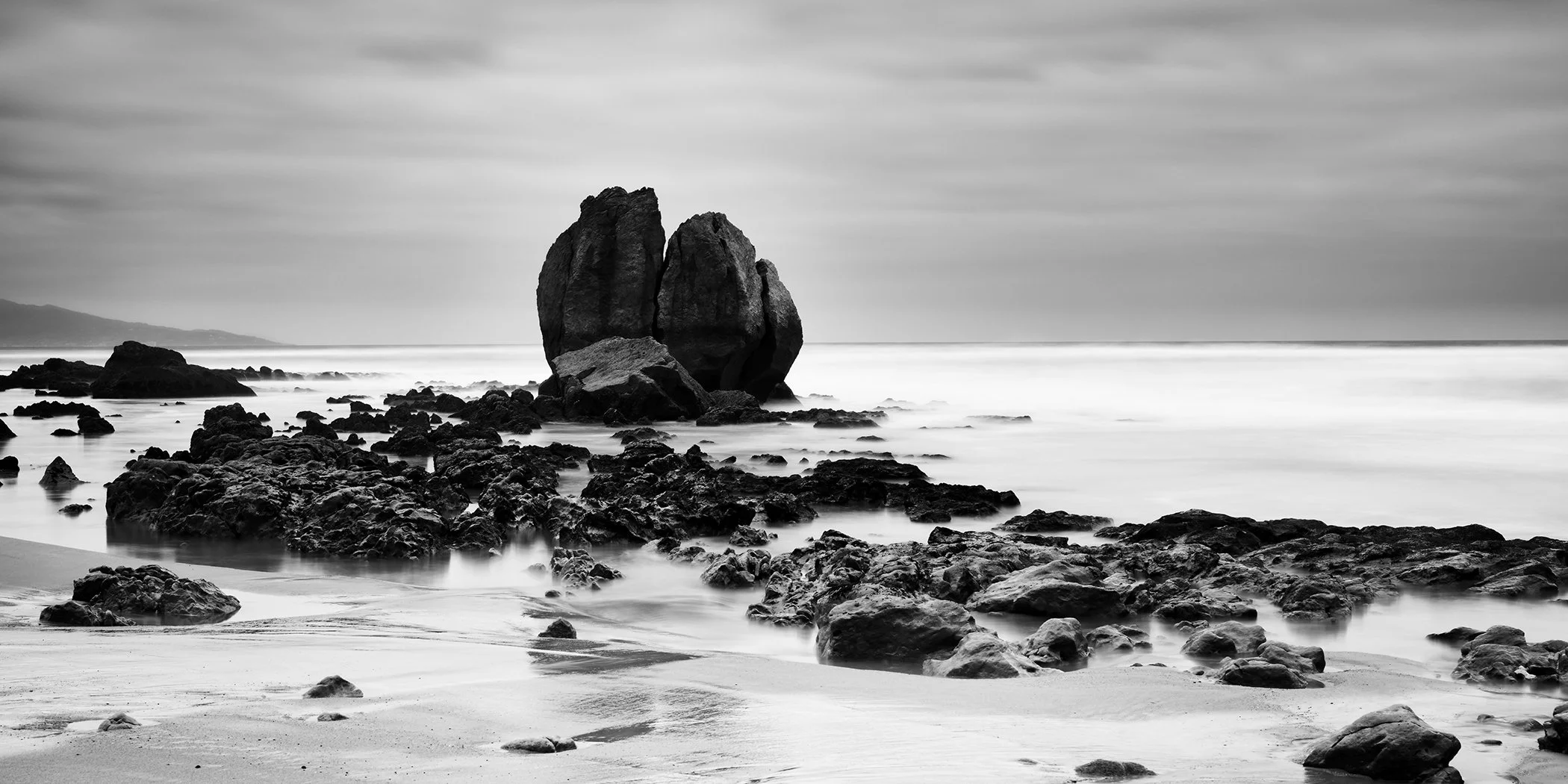 Dramatic monochrome beach scene with large rock formation surrounded by wet sand and waves