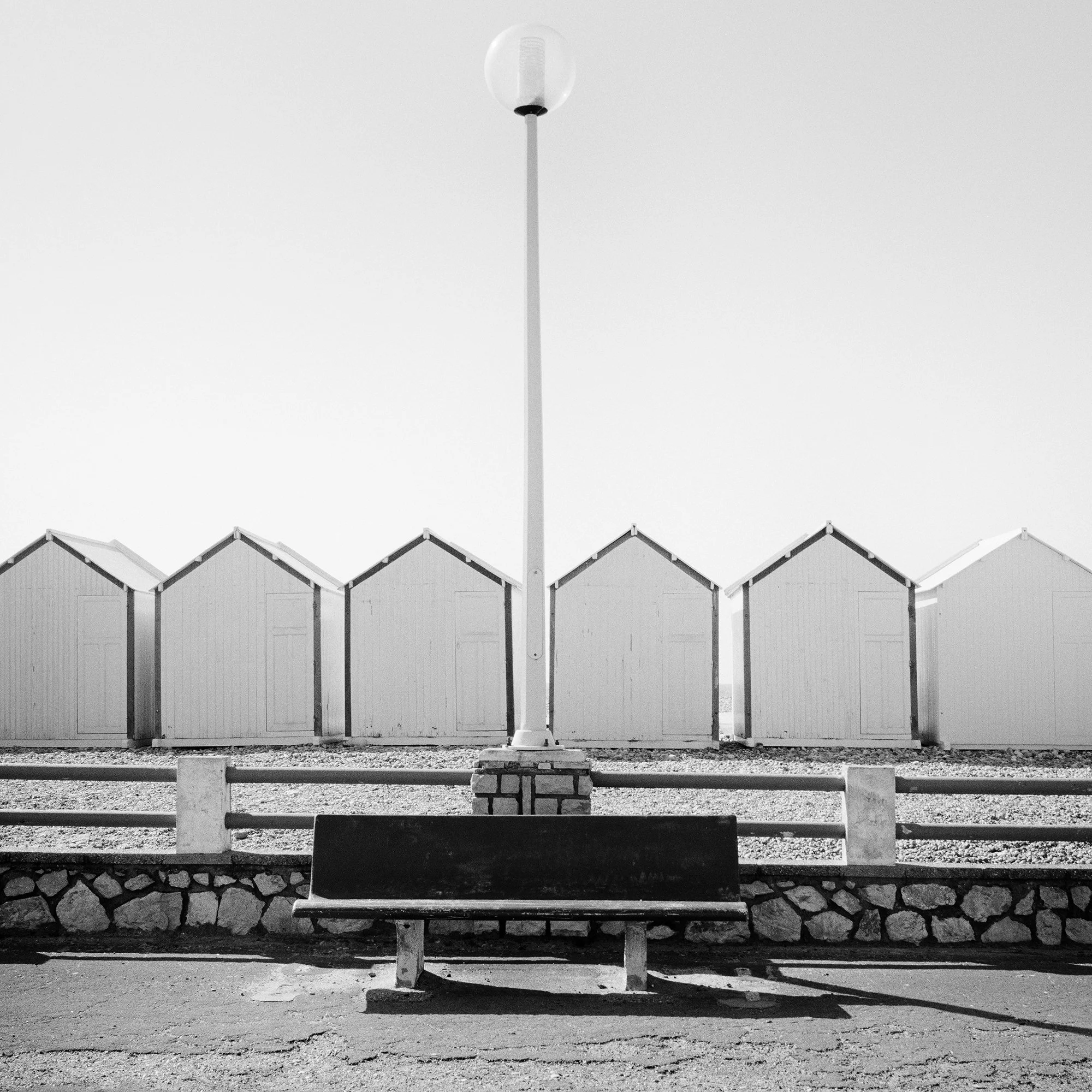 Empty bench on the promenade facing beach huts, centred streetlamp, minimalist black-and-white coastal photograph, Detail 3