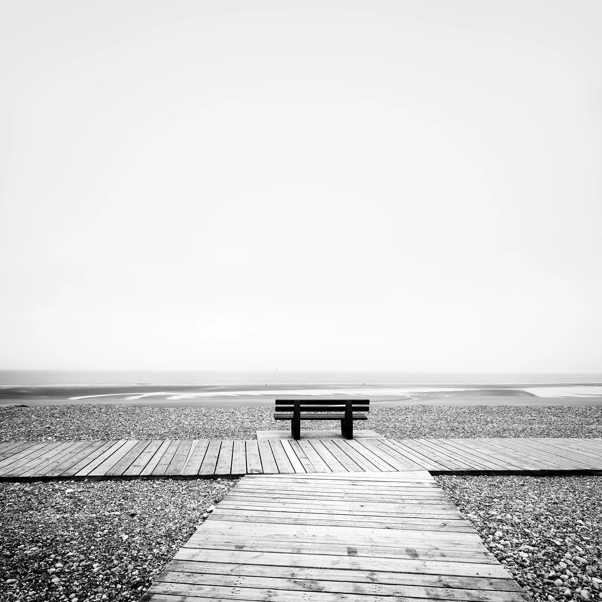 Black-and-white landscape photo of a wooden pathway and bench facing the Atlantic Ocean above a rocky beach