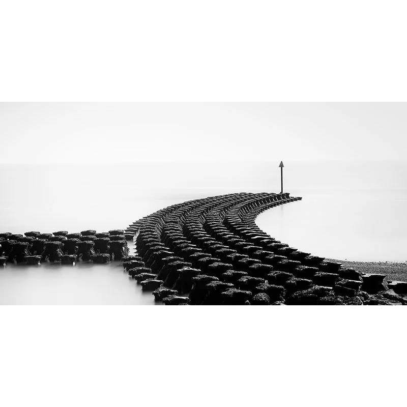 Gerald Berghammer - Black and white minimalist photography. A curved stone jetty extending into a calm body of water, with a tall navigation marker at the end.
