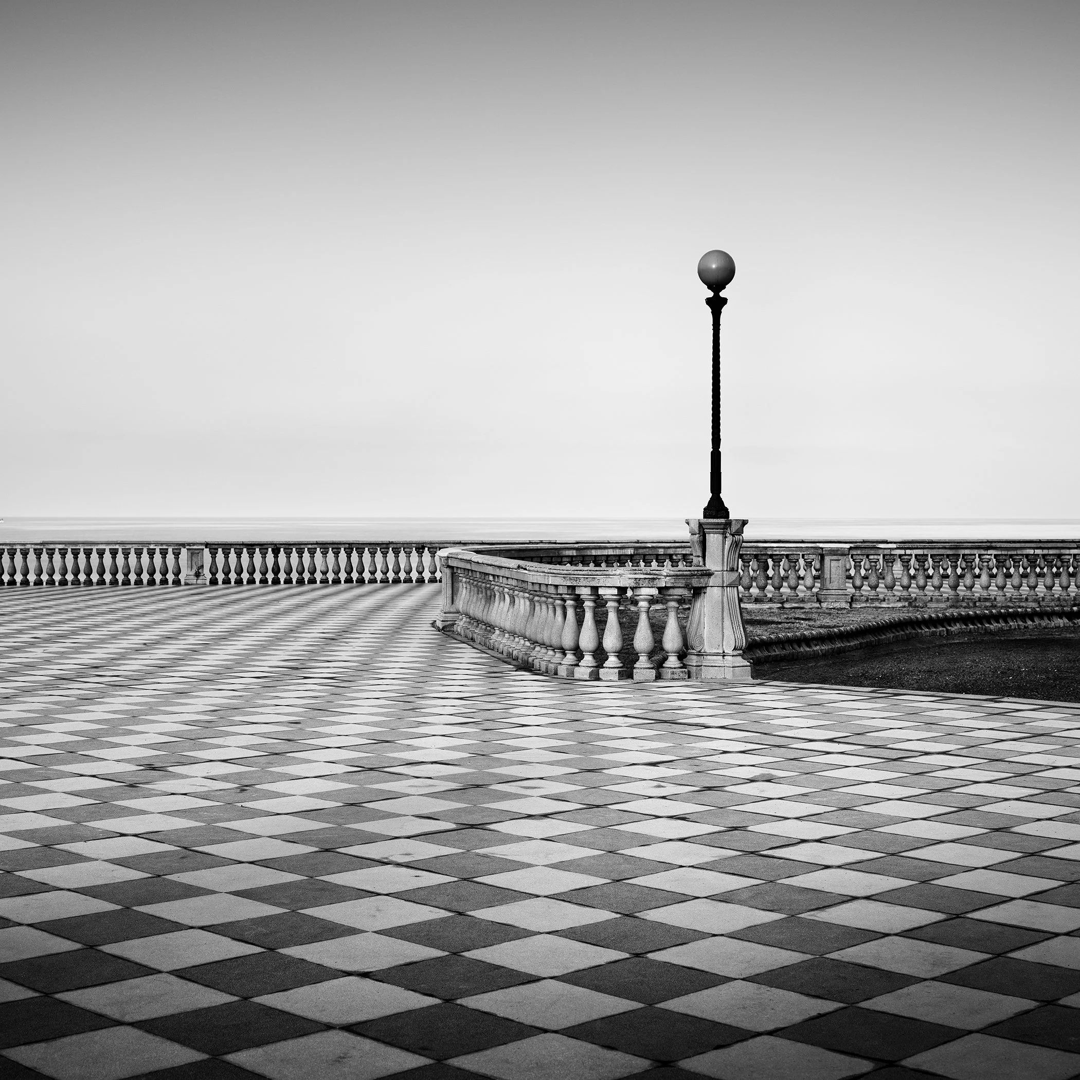 Black-and-white photograph of an empty Tuscan seafront promenade with chequerboard paving and railings leading to the horizon, calm sea beyond.