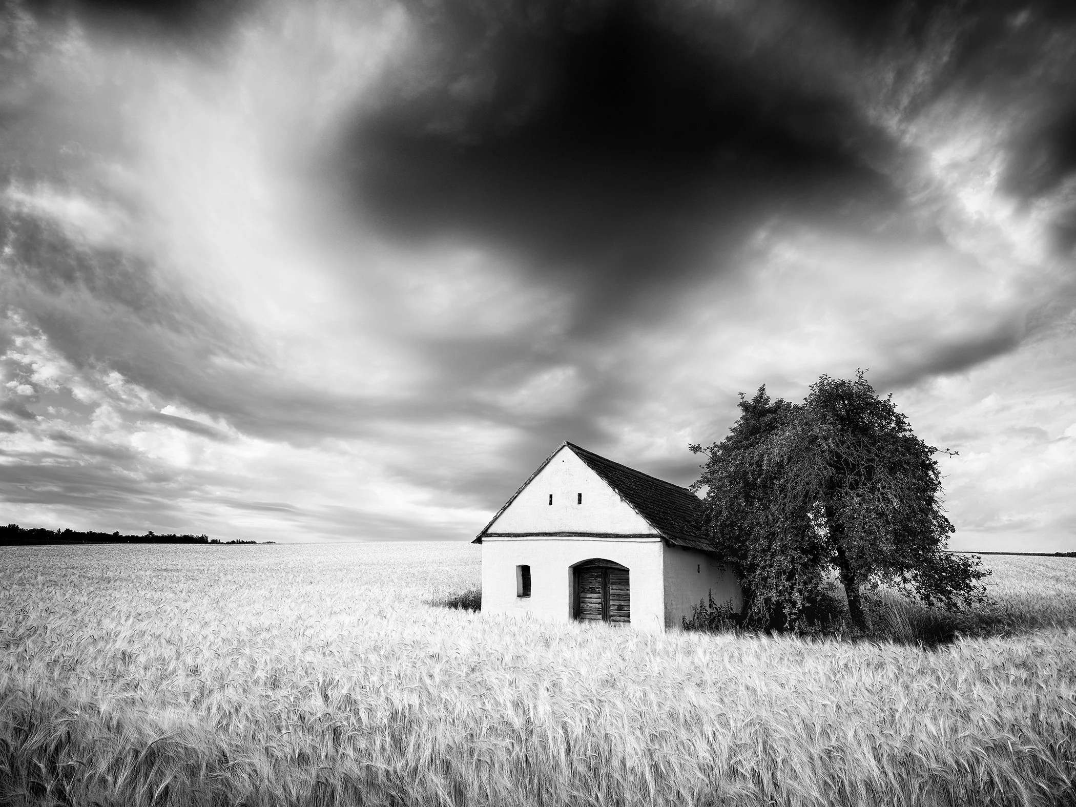 Black-and-white photograph of the Wine Press House isolated in a rippling wheat field, with dark, dramatic storm clouds gathering overhead.