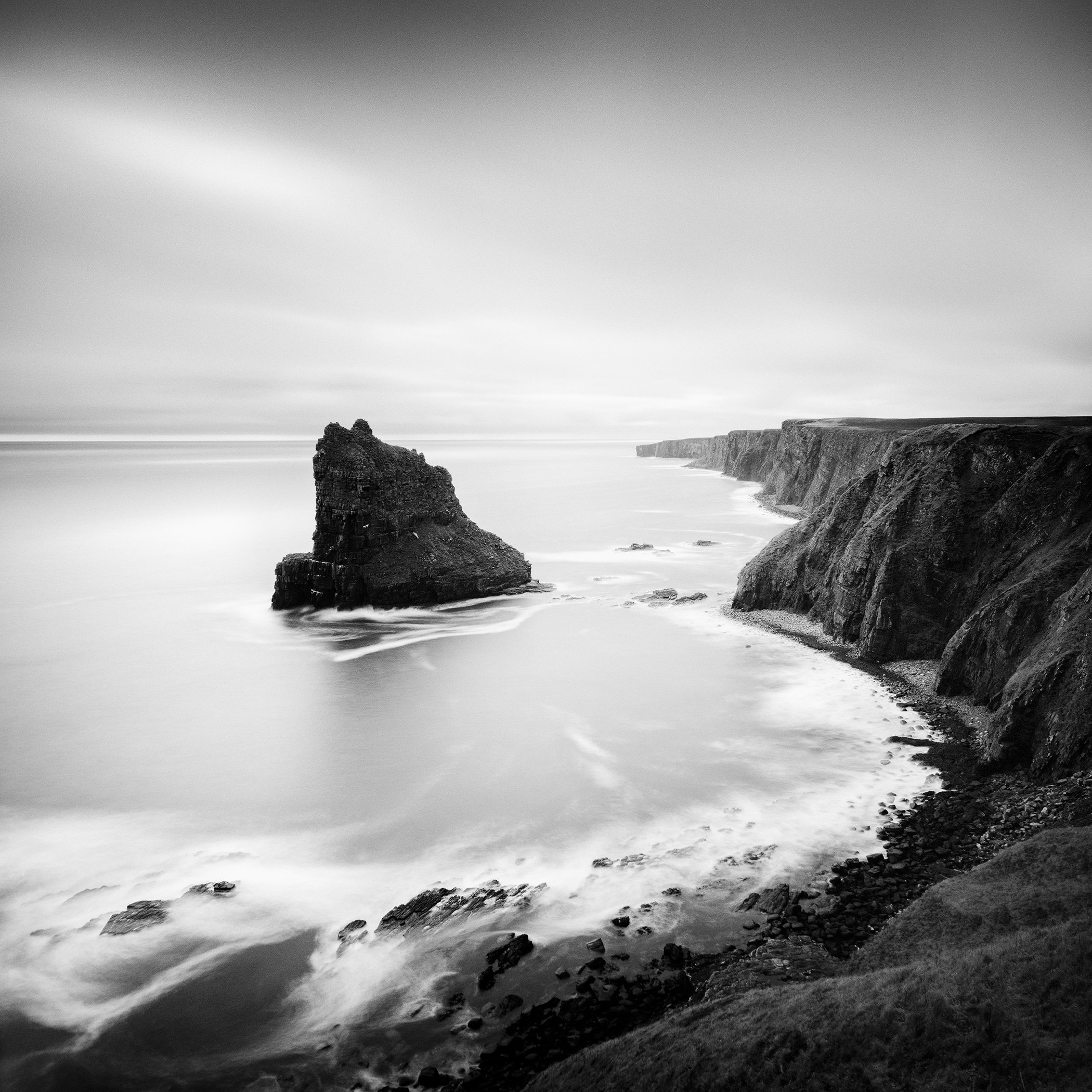 Black and white long exposure photograph of dramatic coastal cliffs and a sea stack on the Scottish coastline