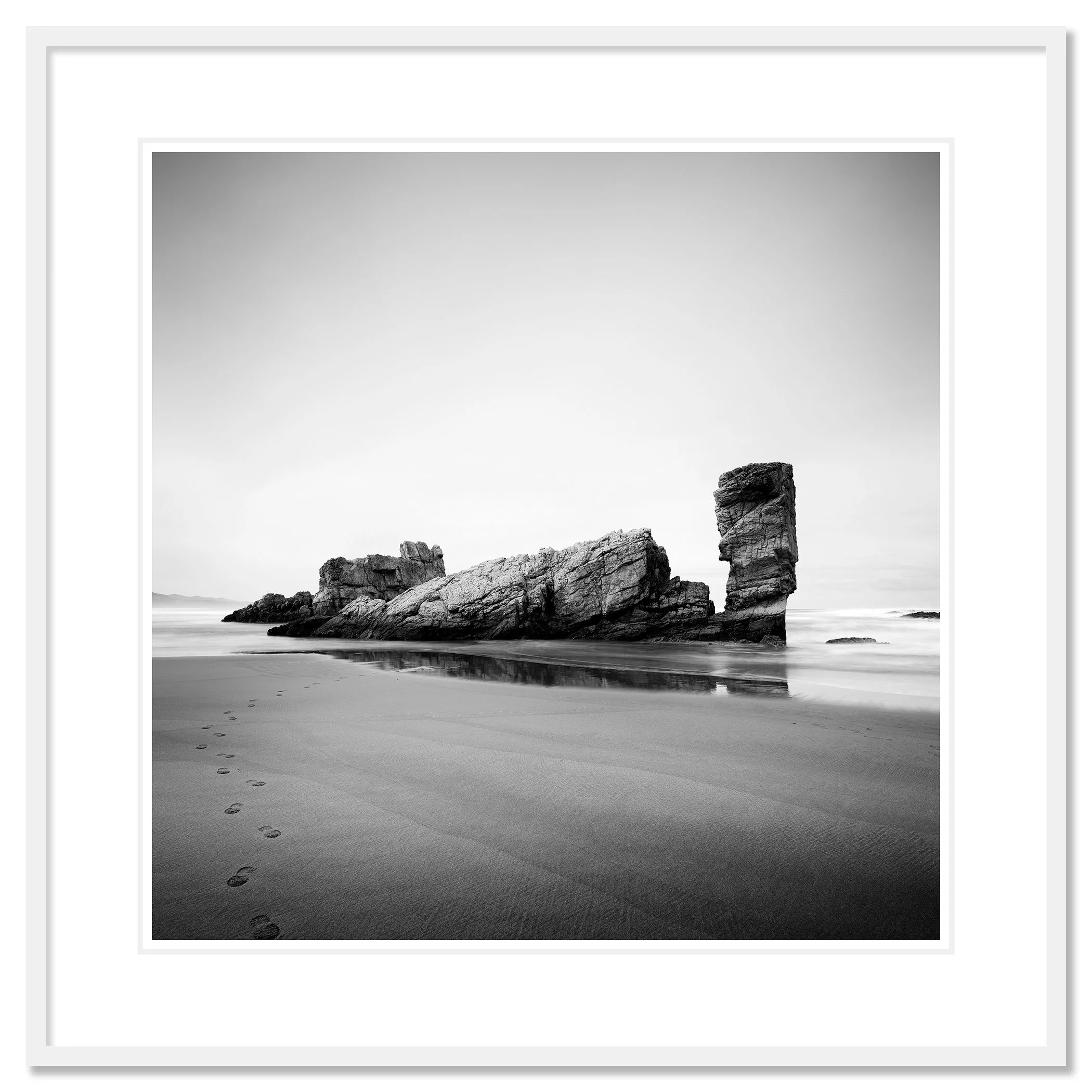 © 2023 Gerald Berghammer. Black-and-white beach scene with wet sand, faint footprints, and a tall rock formation near the shore reflected in shallow water. Classic framed white