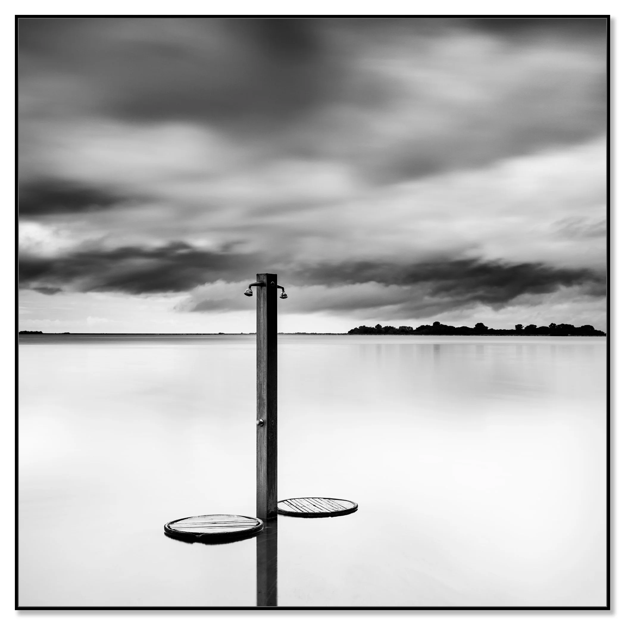 Black-and-white photo of a beach shower standing in calm water under dramatic storm clouds – framed ArtBox black