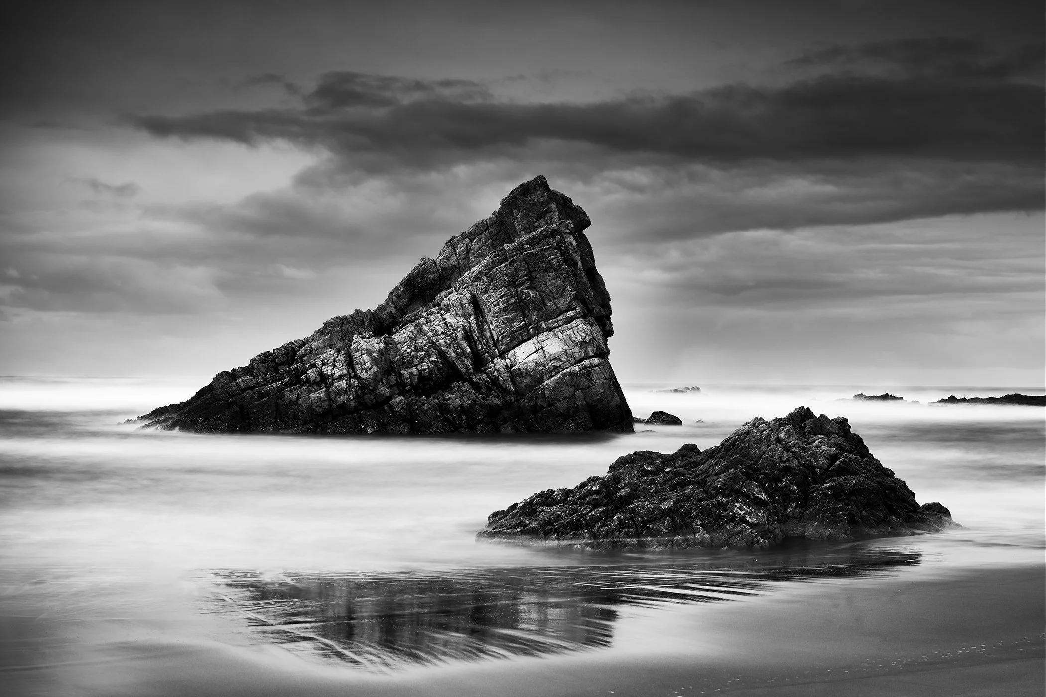 Black-and-white long-exposure seascape of jagged coastal rocks in calm water beneath a cloudy sky, Bay of Biscay, Spain.