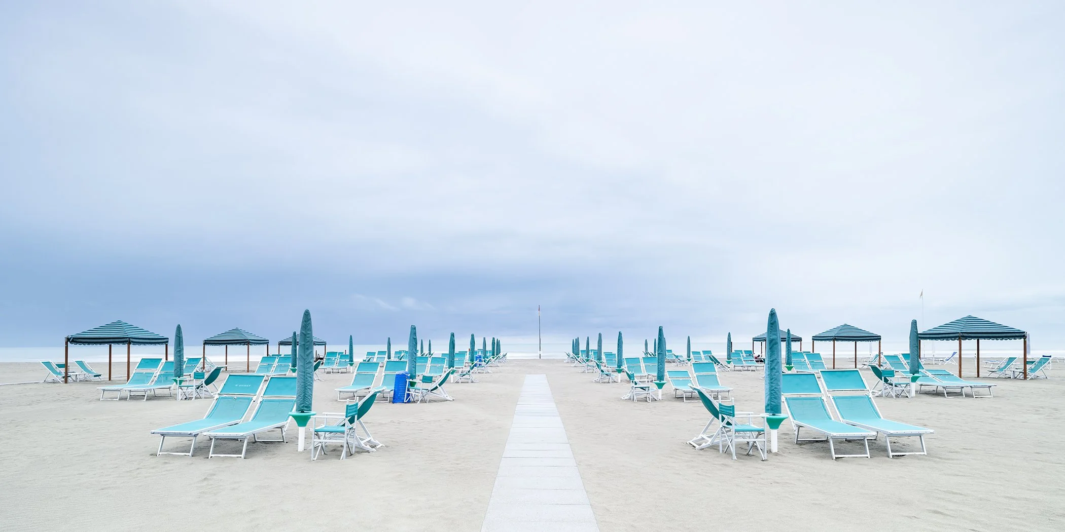 Turquoise sun loungers and parasols arranged symmetrically beside a central walkway on a deserted beach in Italy.