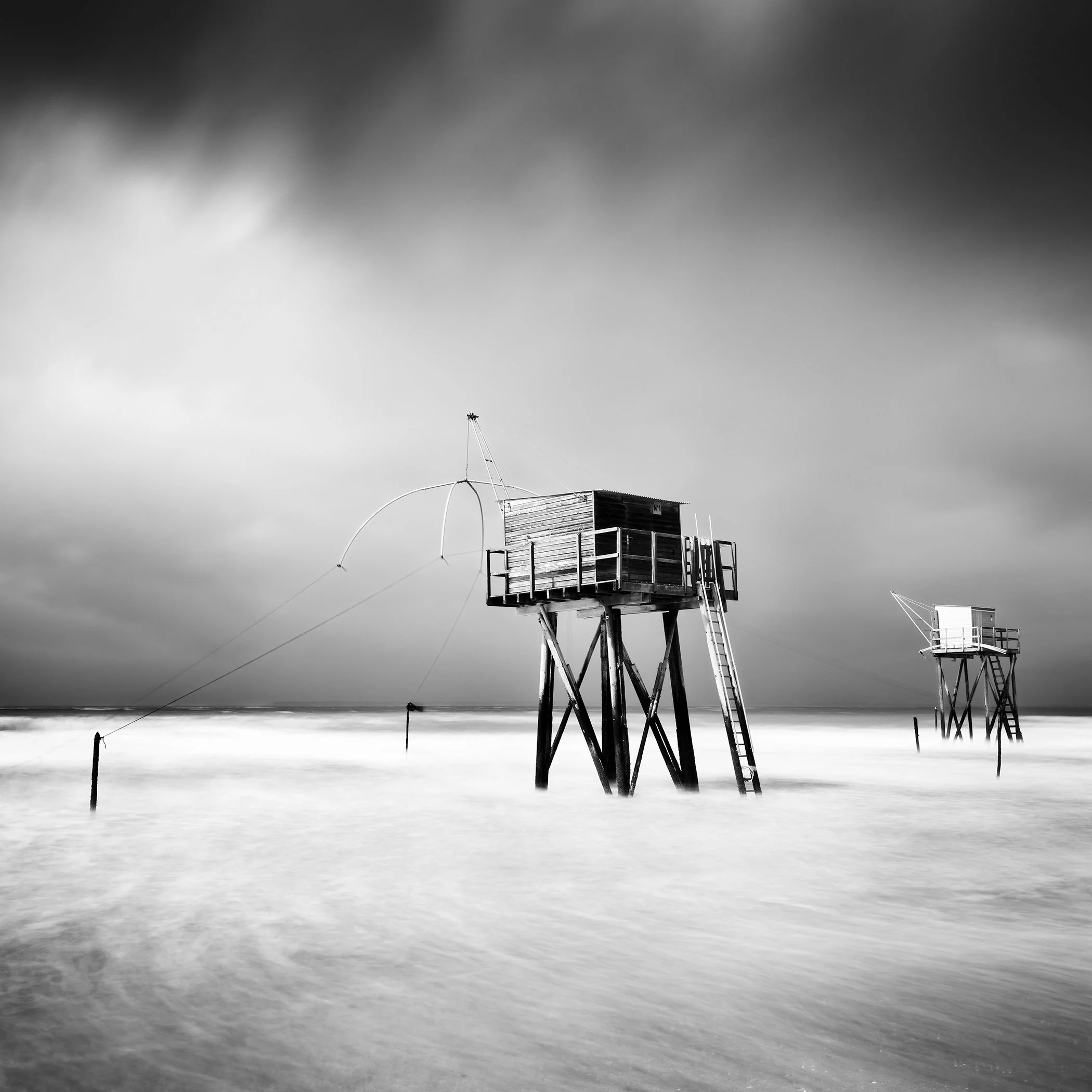 © 2023 Gerald Berghammer - Black-and-white photo of wooden stilt huts in the sea, reached by ladders, with smooth long-exposure water and a dramatic cloudy sky.