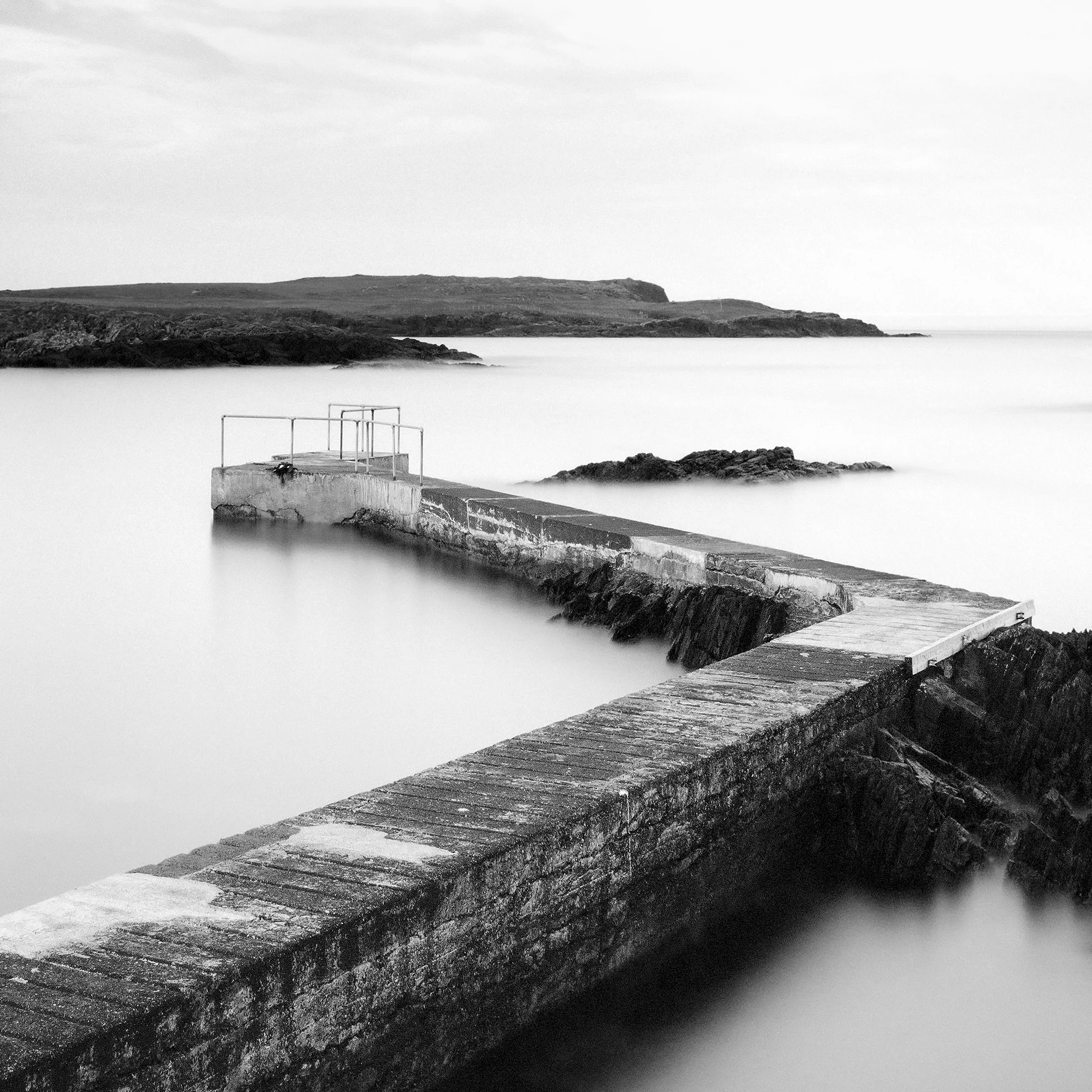 © 2014 Gerald Berghammer - Black and white seascape photography. A Concrete pier extends into calm seawater with rocky edges, under a cloudy sky. Print detail 3