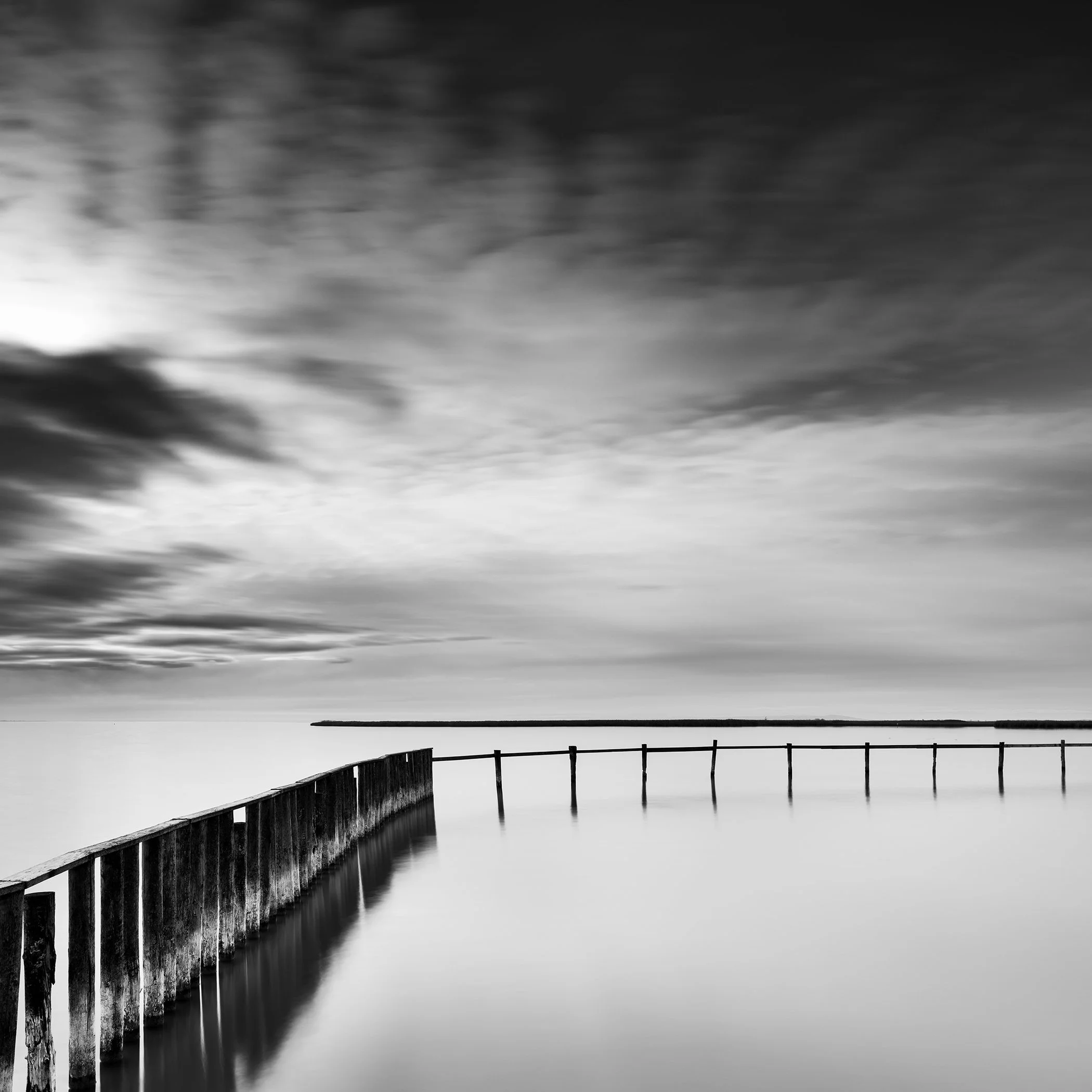 Black and white photograph of a calm lake with a jetty under a dramatic cloudy sky