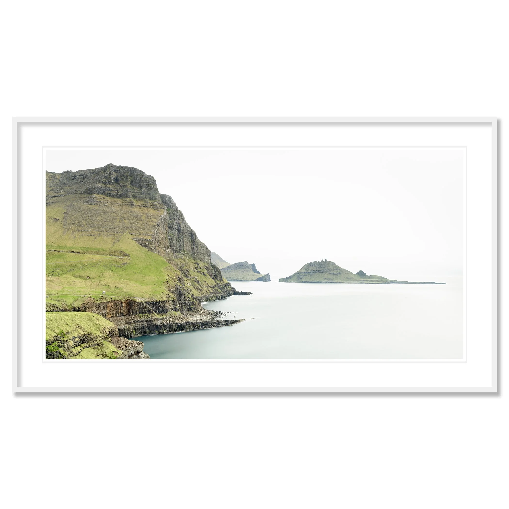 Gerald Berghammer - Color Photography. Cliffs and green hills along a body of water, with distant islands or landmasses in the background under a cloudy sky. Classic framed white