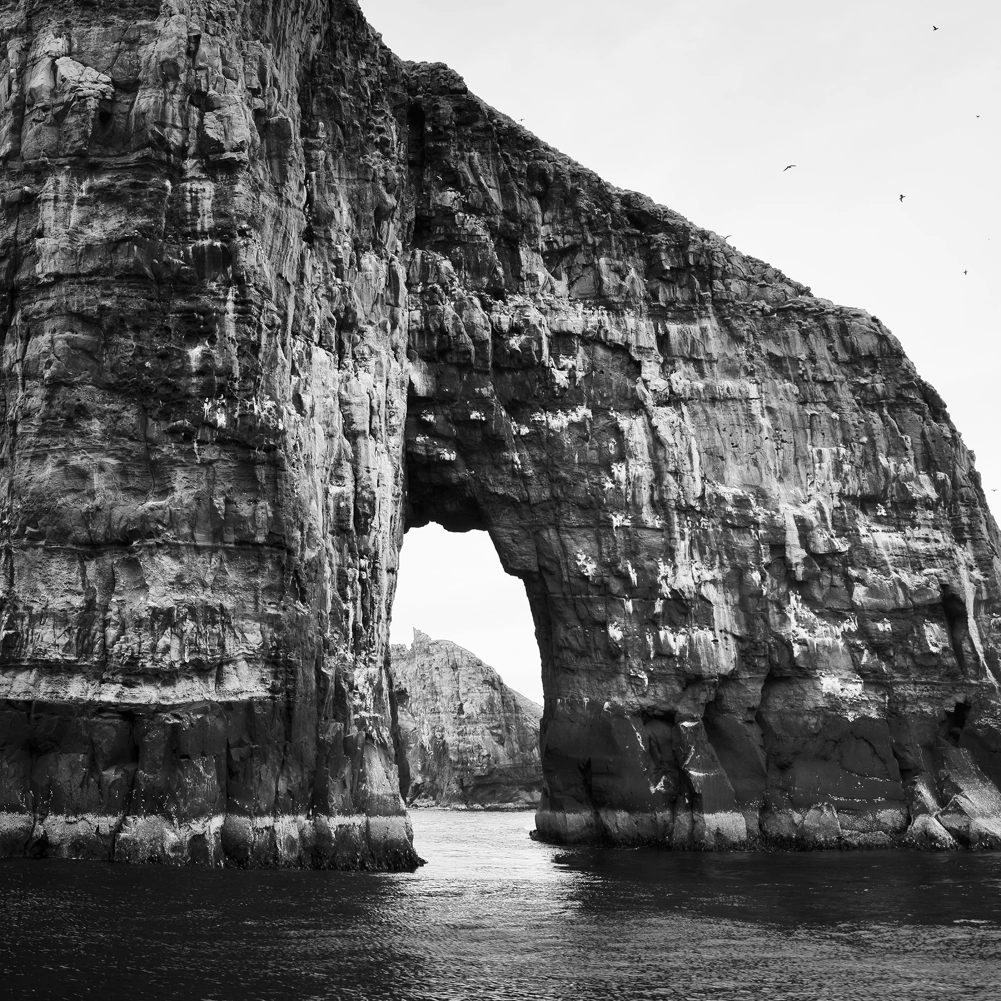 Gerald Berghammer - Black and white seascape photography.  Large natural rock formation with an arch in the middle, surrounded by water and a cloudy sky. Print detail 3