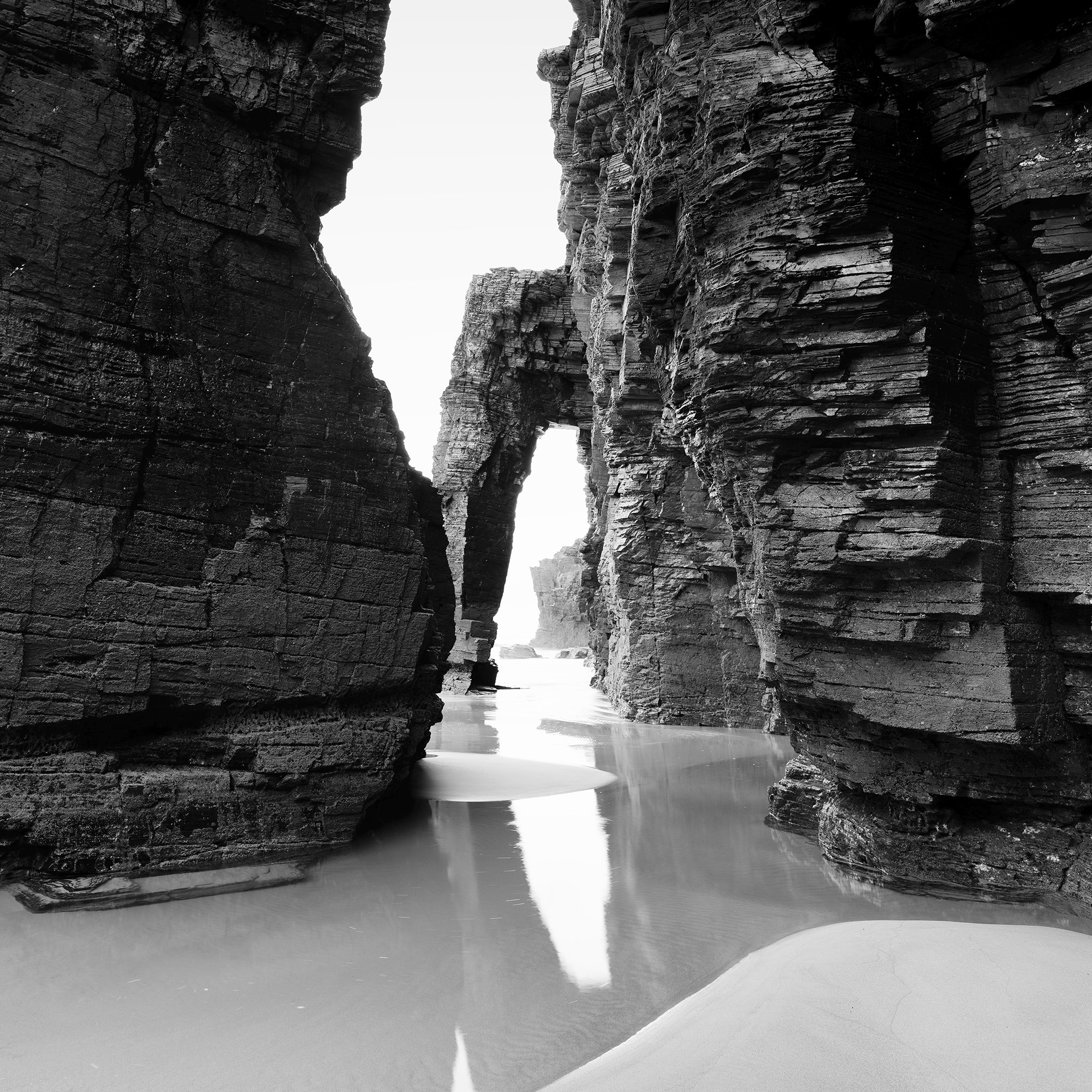 Gerald Berghammer - Black and white beach seascape photography. Large natural rock arch formations on a beach with water reflecting the rocks. Print detail 3