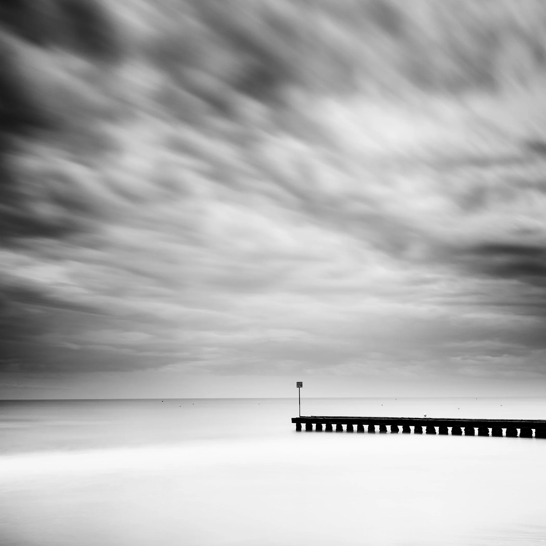 Black and white long exposure seascape with dramatic clouds and a wooden pier extending into calm water