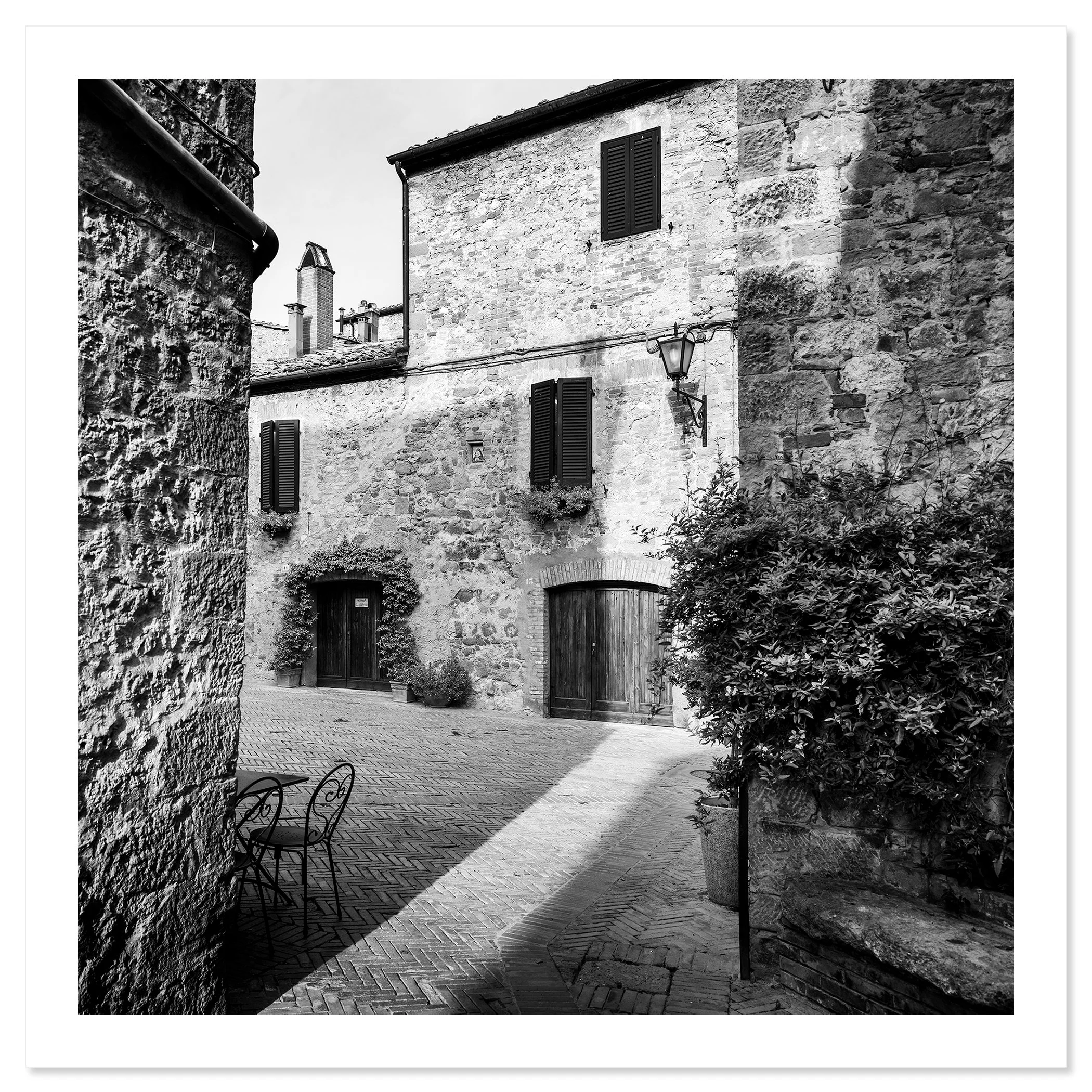 Gerald Berghammer - Black and white cityscape photography. Old stone building with wooden shutters, potted plants and outdoor seating in a narrow courtyard. Fine art print only
