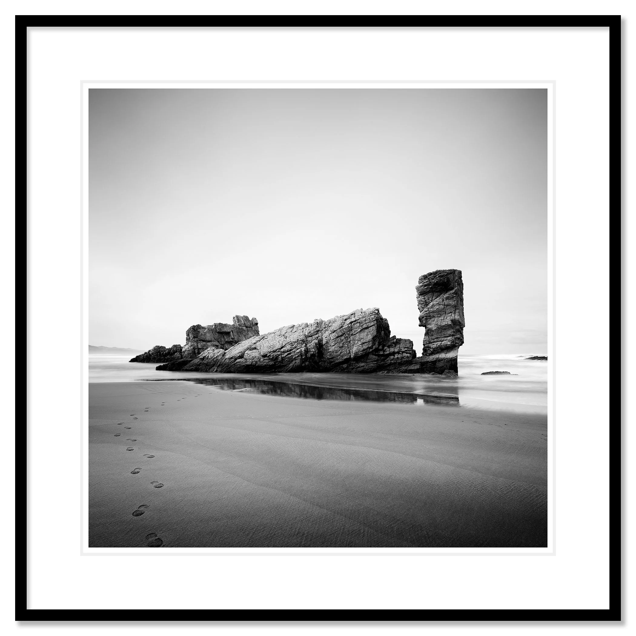 © 2023 Gerald Berghammer. Black-and-white beach scene with wet sand, faint footprints, and a tall rock formation near the shore reflected in shallow water. Classic framed black