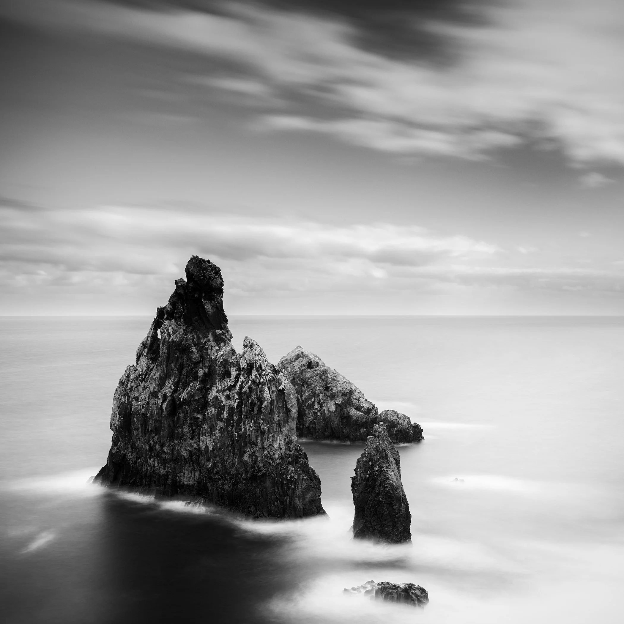 Black-and-white long-exposure seascape of rugged rocks rising from a calm ocean, with silky-smooth water and dramatic clouds overhead.