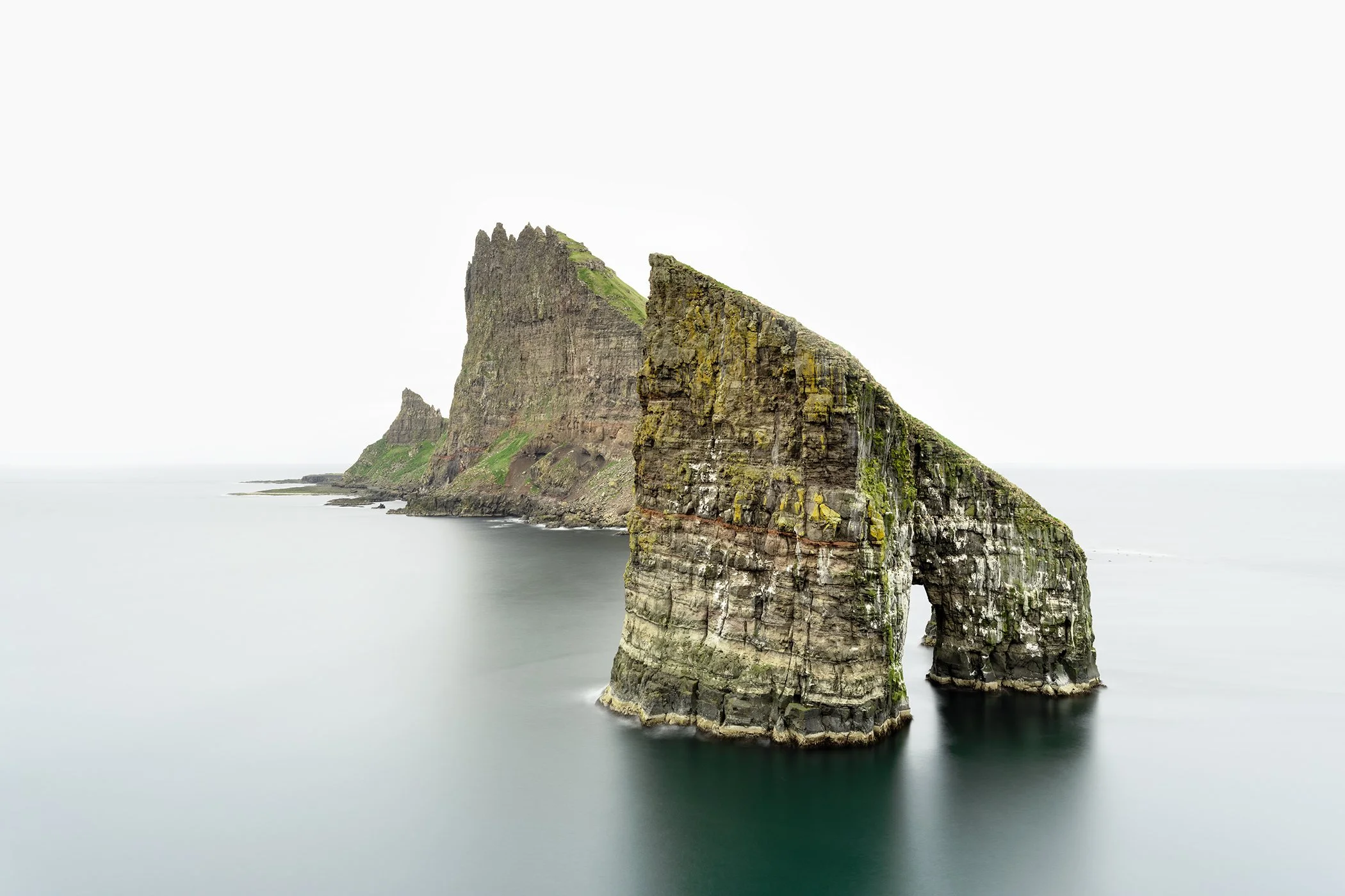 Rugged Drangarnir sea stack with natural rock arch rising from calm misty water, Faroe Islands