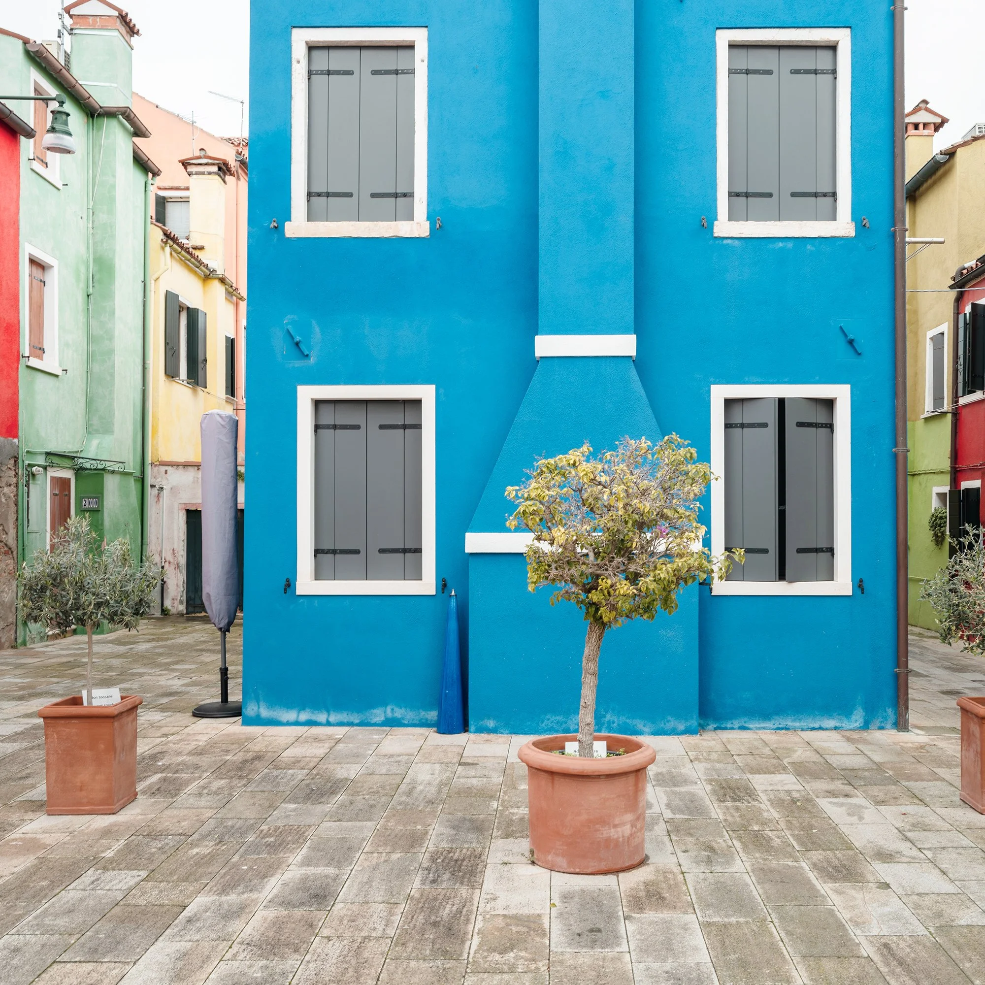 © 2025 Gerald Berghammer - Color cityscape photo. Colorful row houses with a prominent blue house in the center, potted small trees in front, and a paved courtyard. Print detail 1