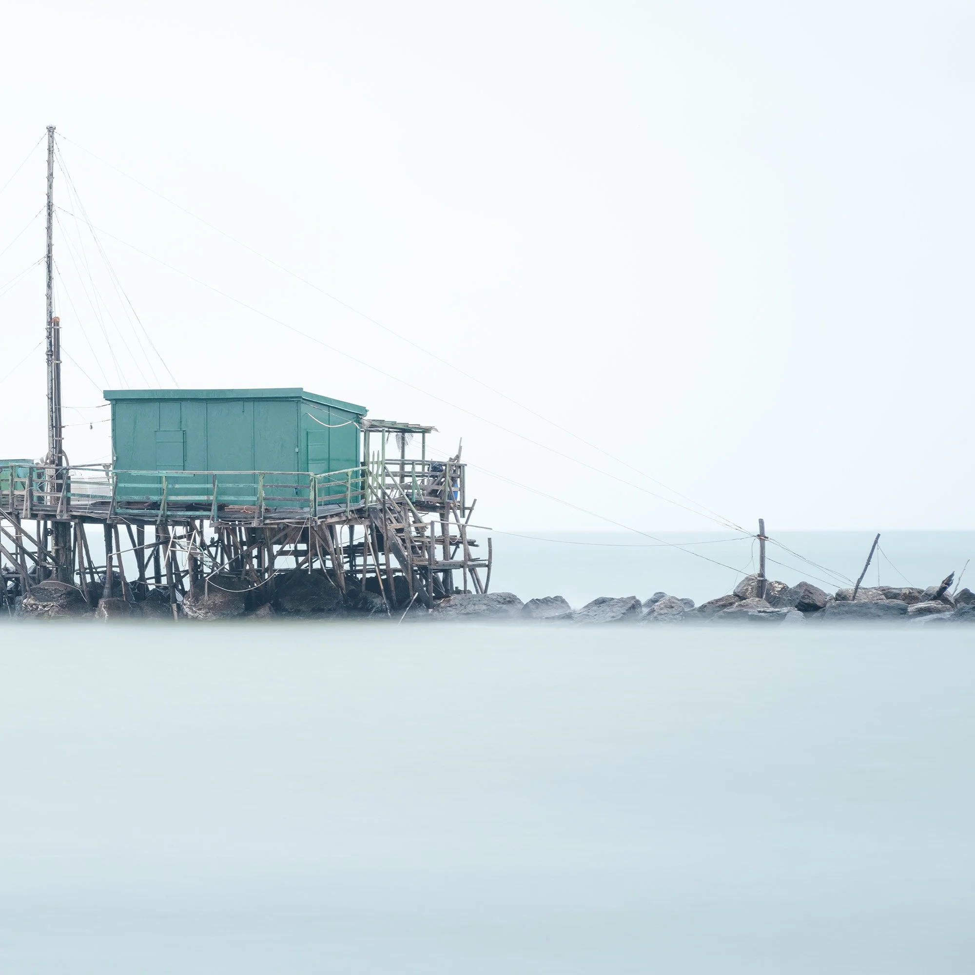 © 2024 Gerald Berghammer - Color minimalist photography. A small house built on stilts over water on rocks, with a fishing pole, in a minimalist, misty seascape. Print detail 2