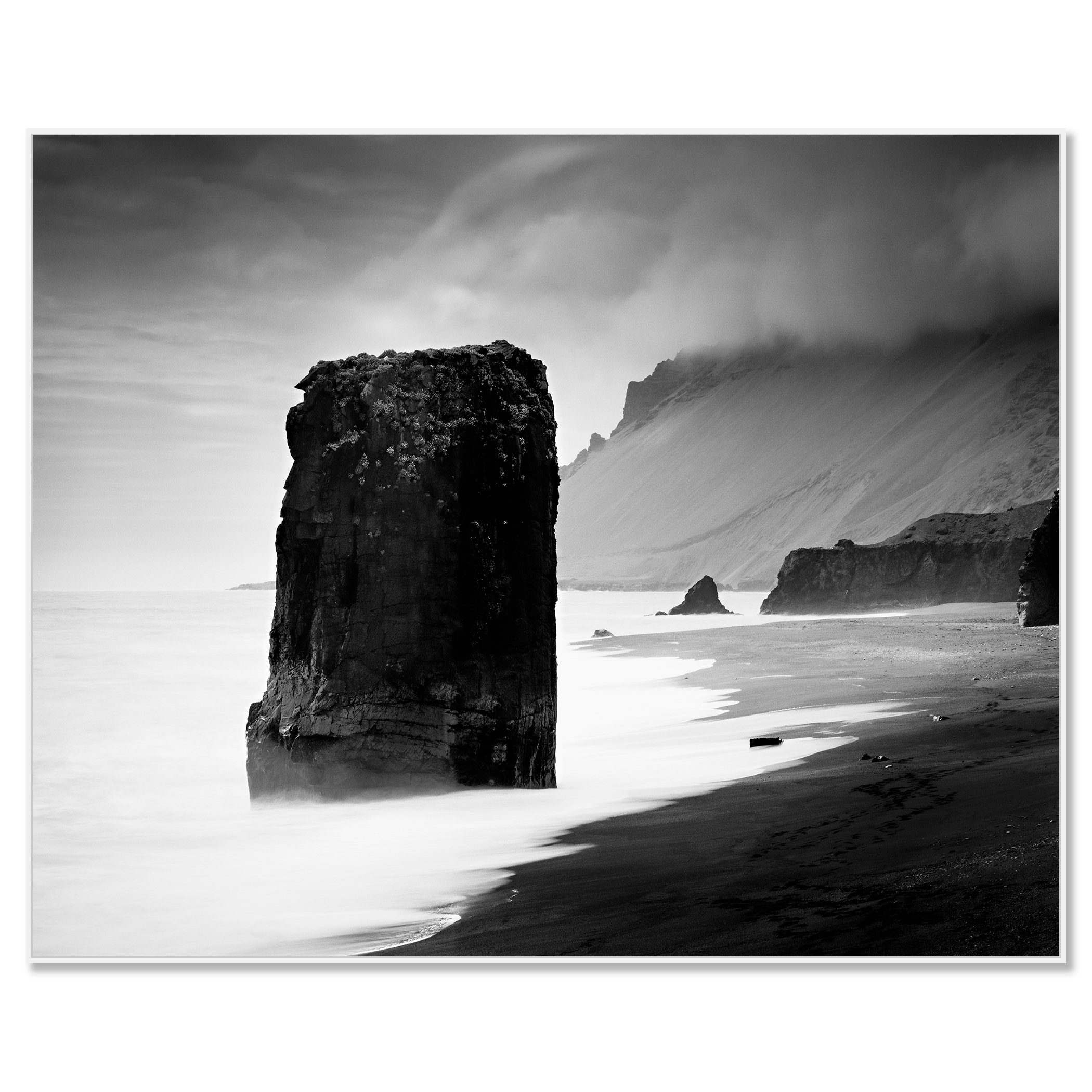 Black-and-white photograph of a rock monolith in the sea, with smooth long-exposure water, dark sand, and misty cliffs, framed ArtBox white