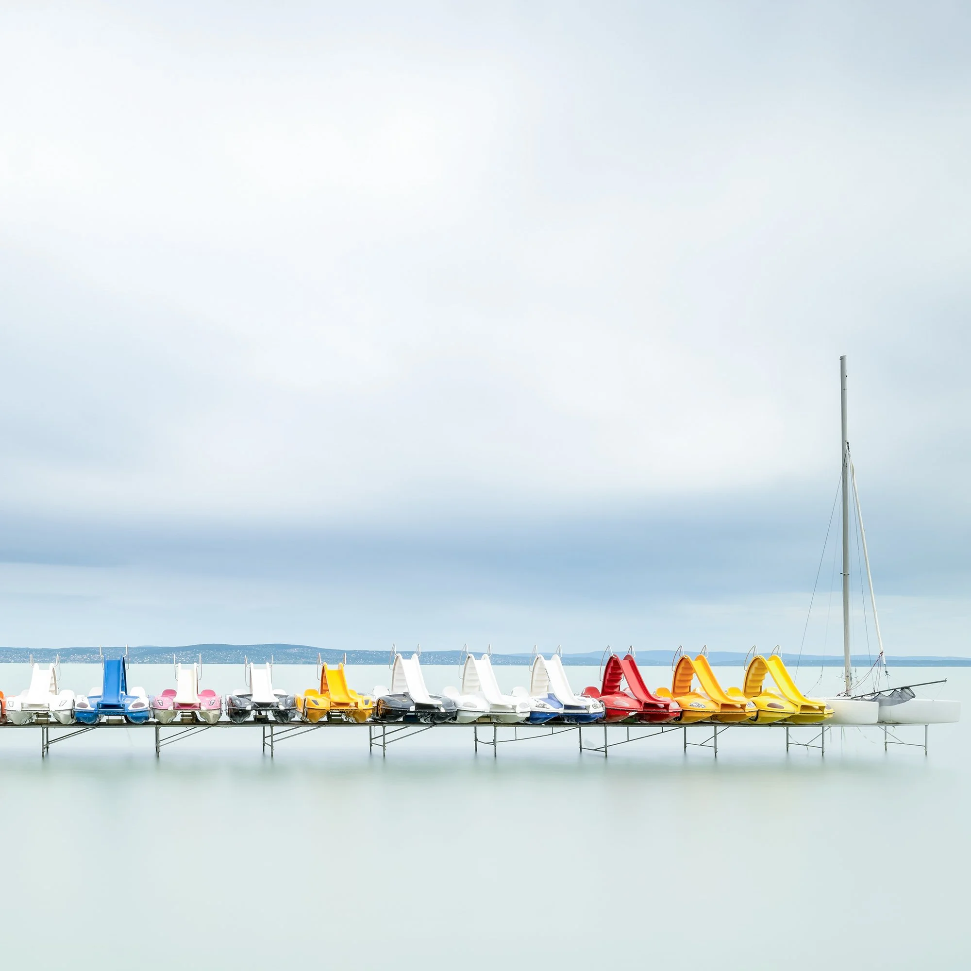 Gerald Berghammer - Color waterscape photography. Colorful pedal boats and a catamaran lined up on a dock on a calm body of water under a cloudy sky. Print detail 2