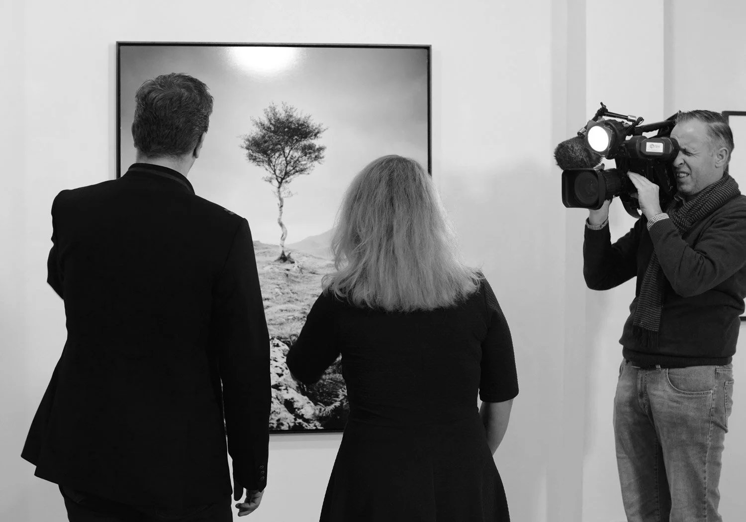 Cameraman filming Gerald Berghammer and Ina Forstinger in front of a large framed black-and-white photograph of a solitary tree by Gerald Berghammer during ESSENTIAL LIGHT 2018 at Galerie STP in Greifswald.