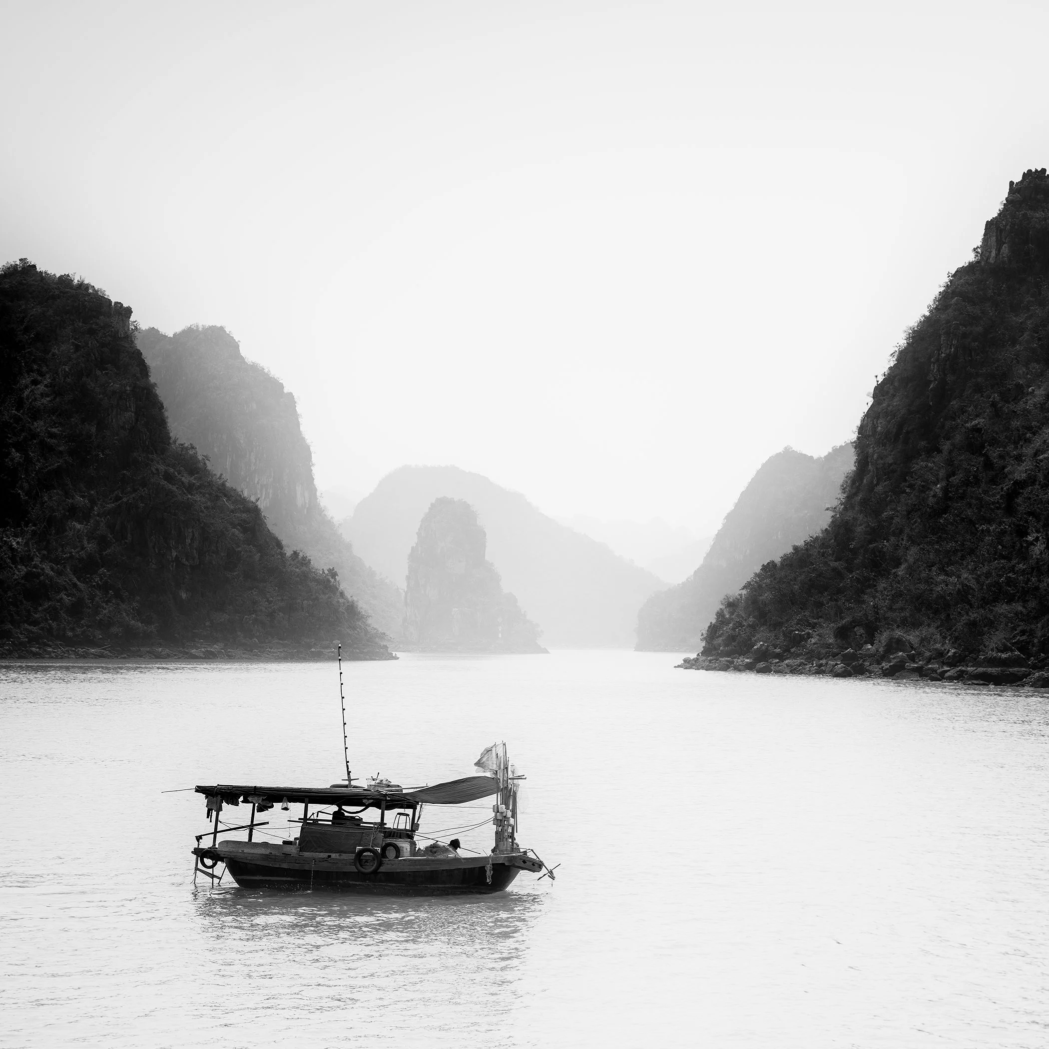 Small fishing boat on calm water among misty limestone karsts in Ha Long Bay, Vietnam, photographed in black and white by Gerald Berghammer.