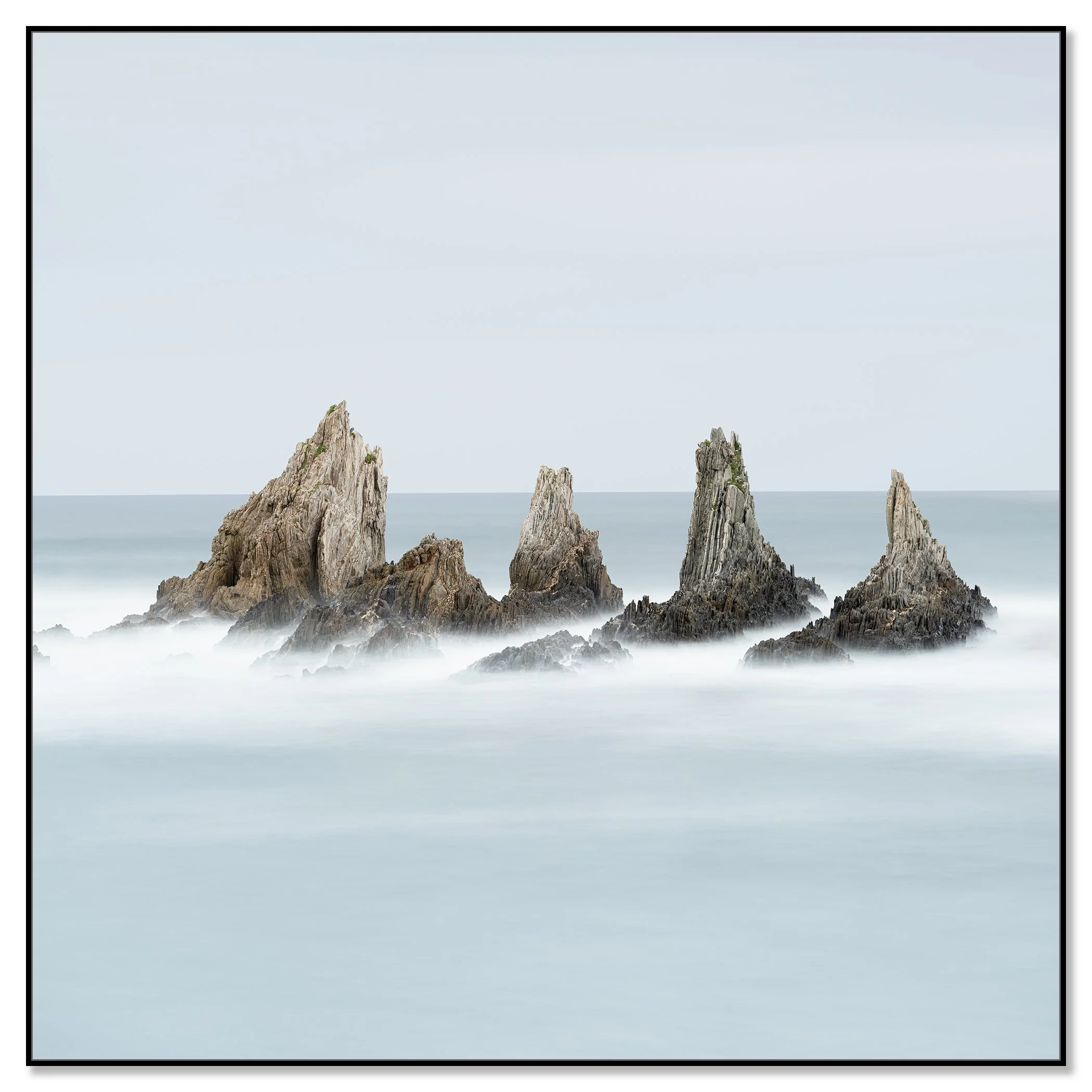 Gerald Berghammer - Color long exposure fine art photography. A seascape with five tall, jagged rocks emerging from the water and a hazy, blue sky. Chromaluxe framed black