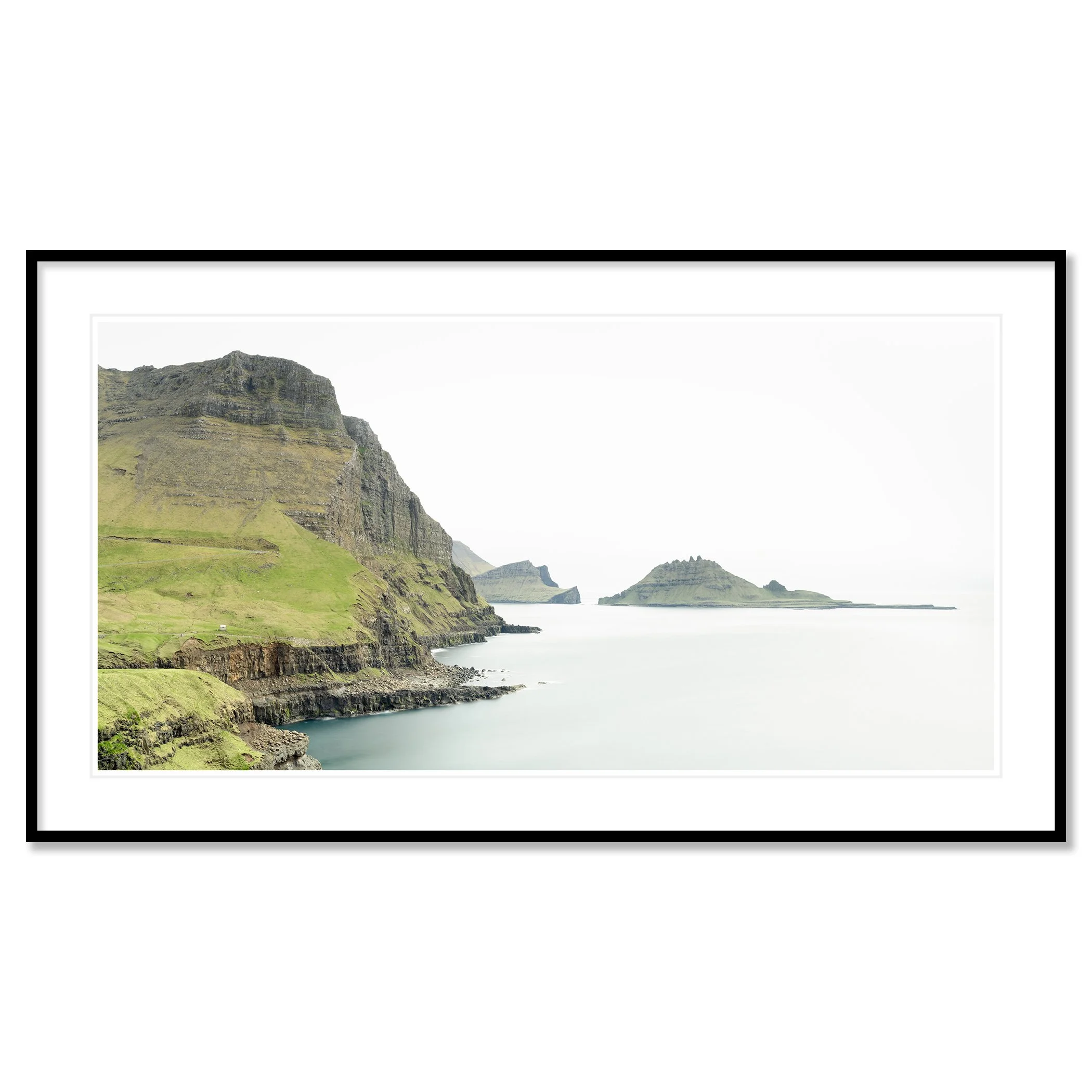 Gerald Berghammer - Color Photography. Cliffs and green hills along a body of water, with distant islands or landmasses in the background under a cloudy sky. Classic framed black