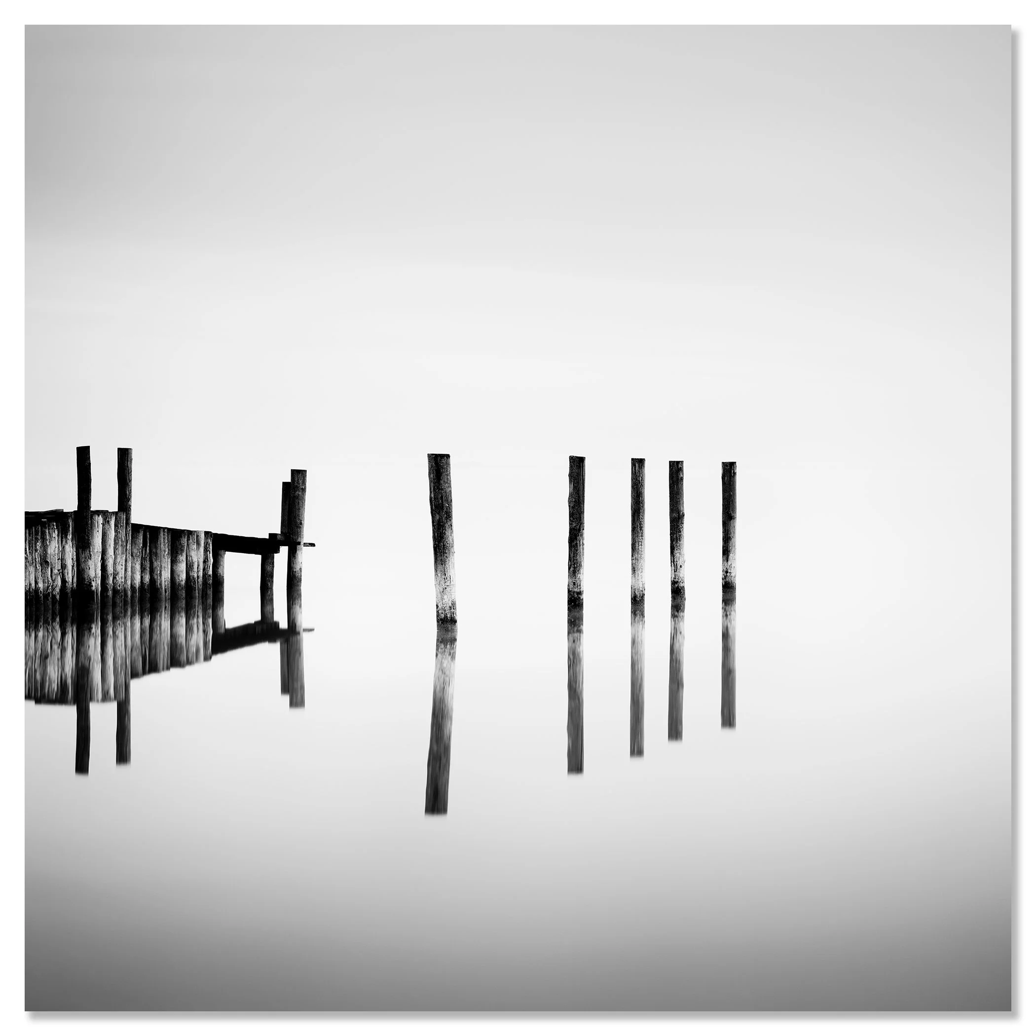 Black-and-white photo of weathered posts and dock reflected in calm lake water beneath an overcast sky – dibond frameless