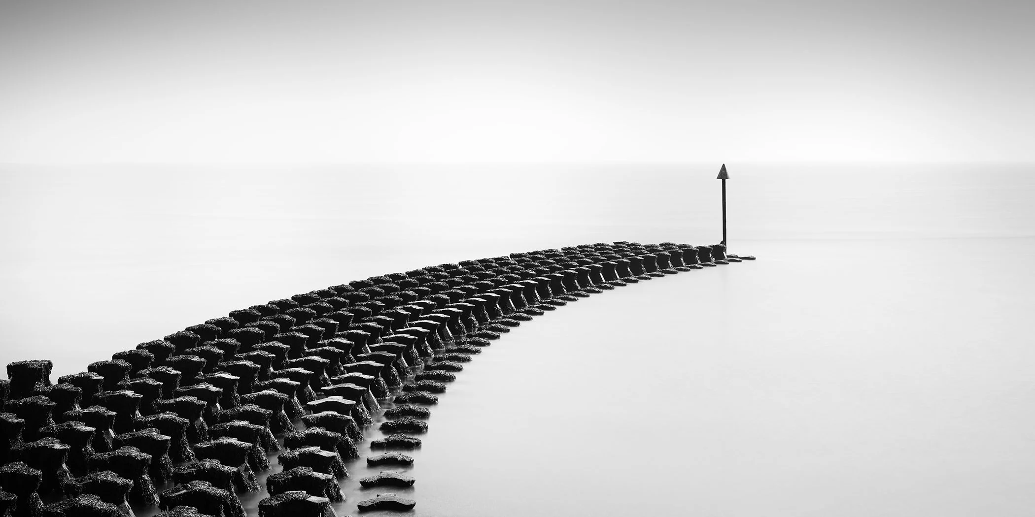 © 2025 Gerald Berghammer. Curved stone breakwater extending into calm water in a minimalist black and white seascape with a navigation marker at the horizon.