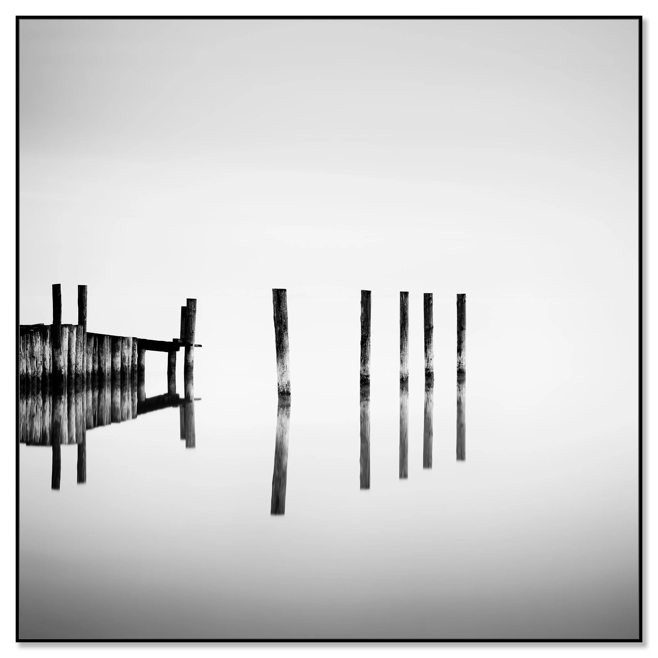 Black-and-white photo of weathered posts and dock reflected in calm lake water beneath an overcast sky – framed ArtBox black