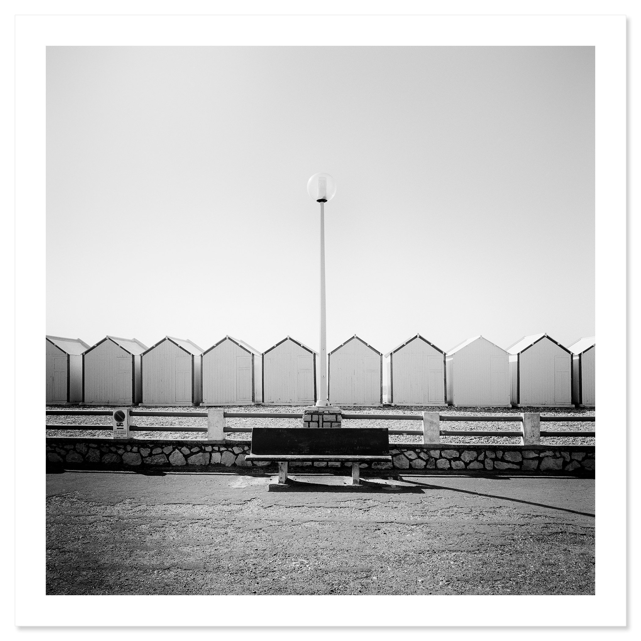 Empty bench on the promenade facing beach huts, centred streetlamp, minimalist black-and-white coastal photograph, Print only