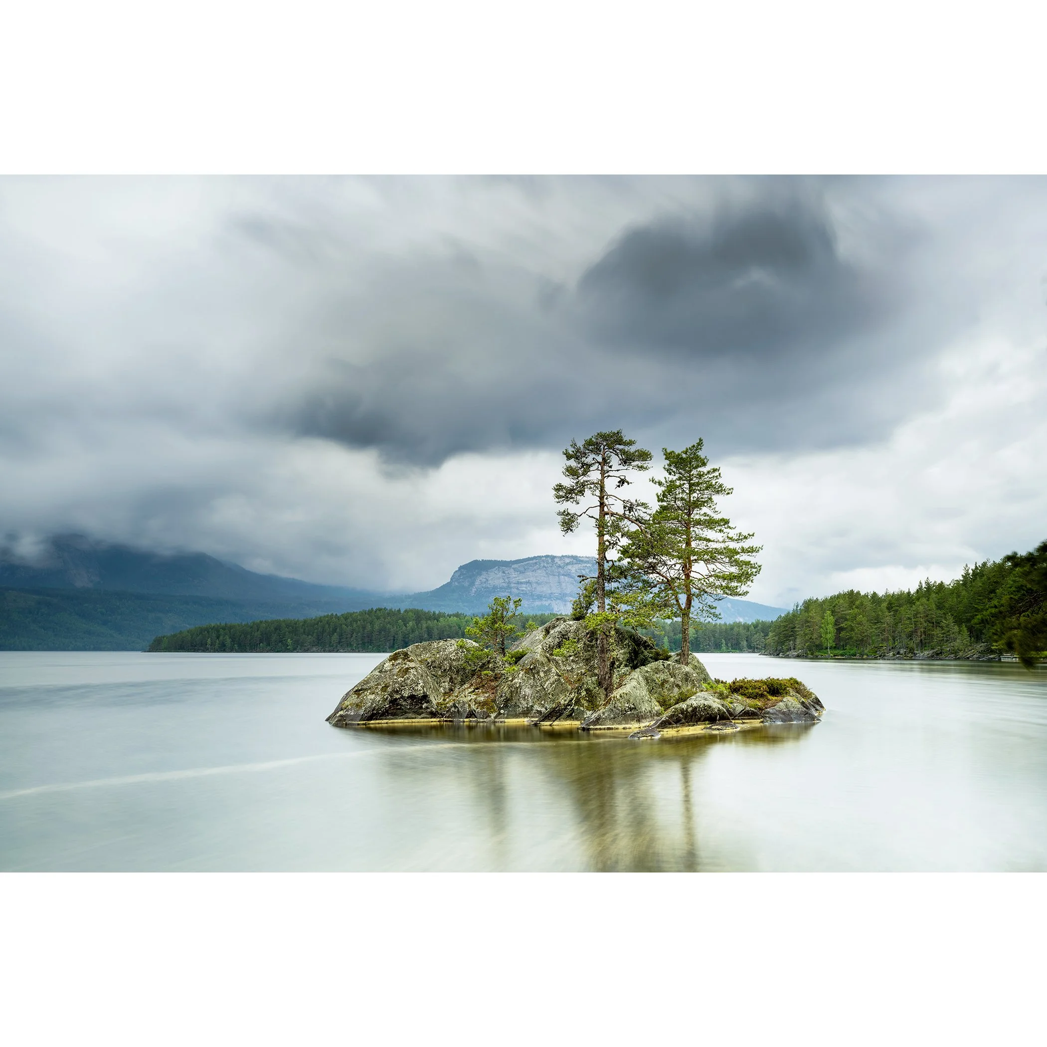 Gerald Berghammer - Color Photography. A small rocky island with tall pine trees surrounded by a calm lake under a cloudy sky and mountains behind.