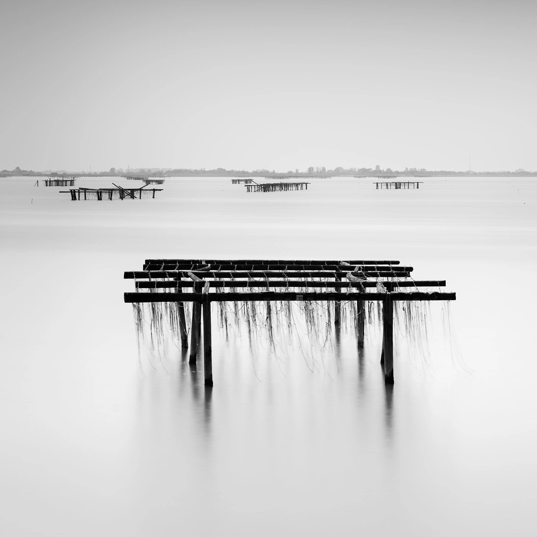 Serene black-and-white coastal scene with wooden structures in smooth long-exposure water and faint reflections below.