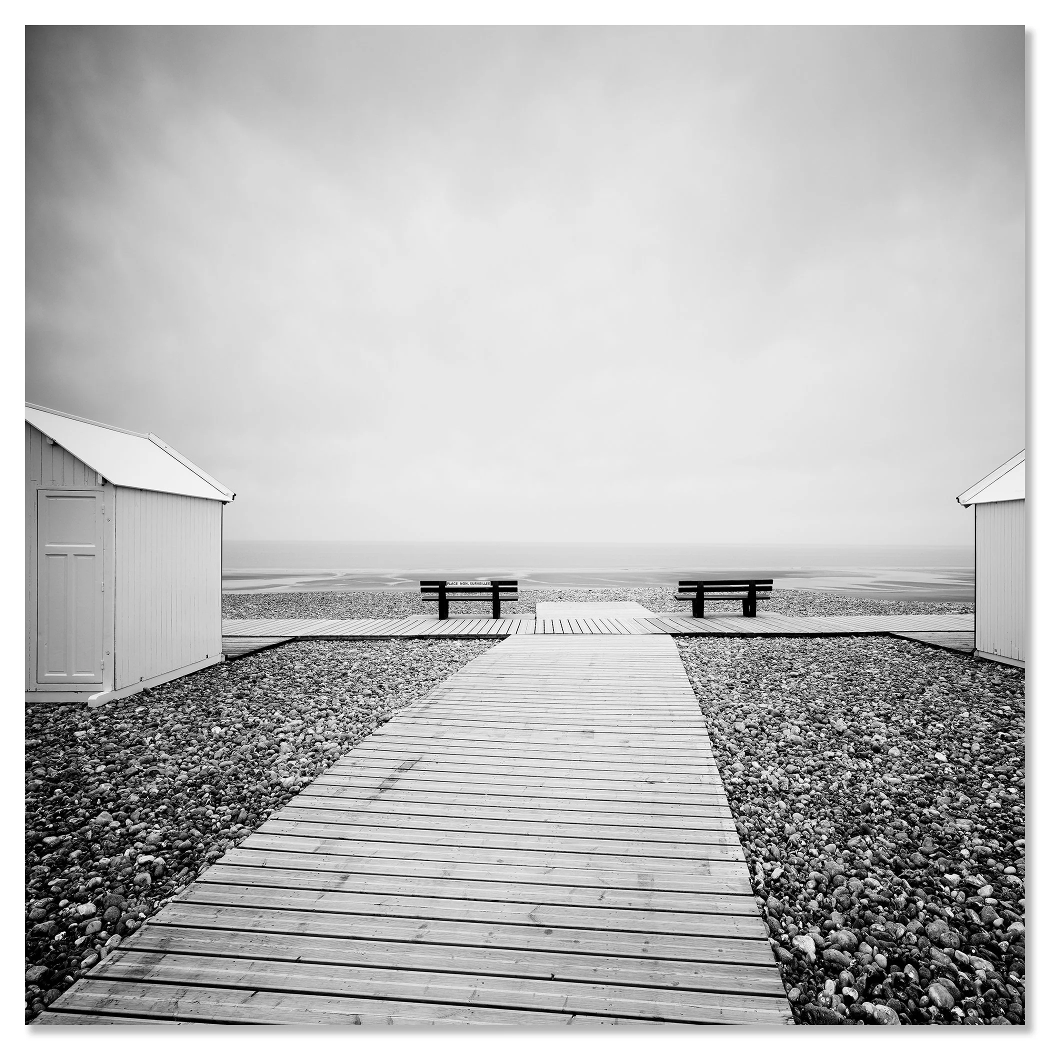 Black and white photo of a wooden boardwalk leading across a pebble beach to benches facing the sea – dibond frameless