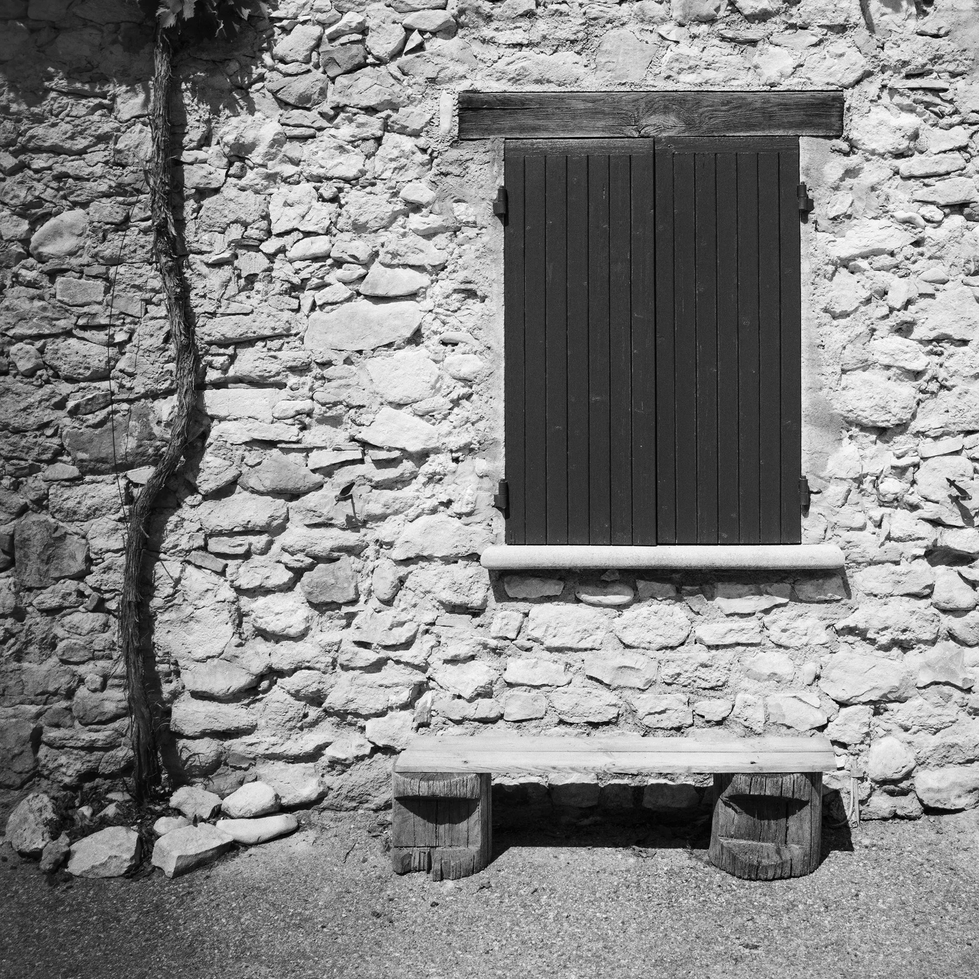 Gerald Berghammer - Black & white photography. Stone wall with a closed wooden window shutter, flanked by climbing vines on both sides, and a small bench. Print detail 1