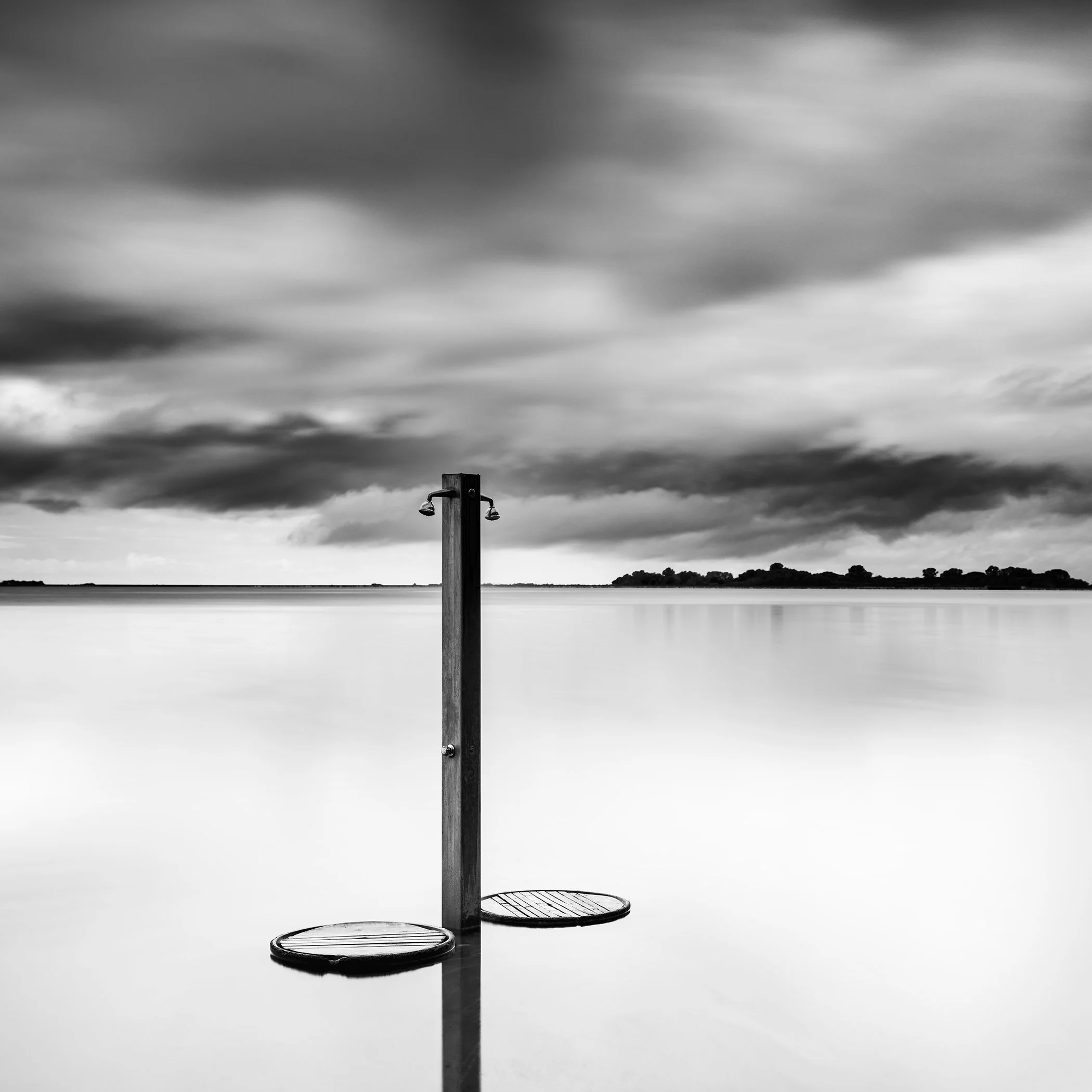 Minimalist black-and-white landscape with a beach shower pole in still water and dark clouds above.