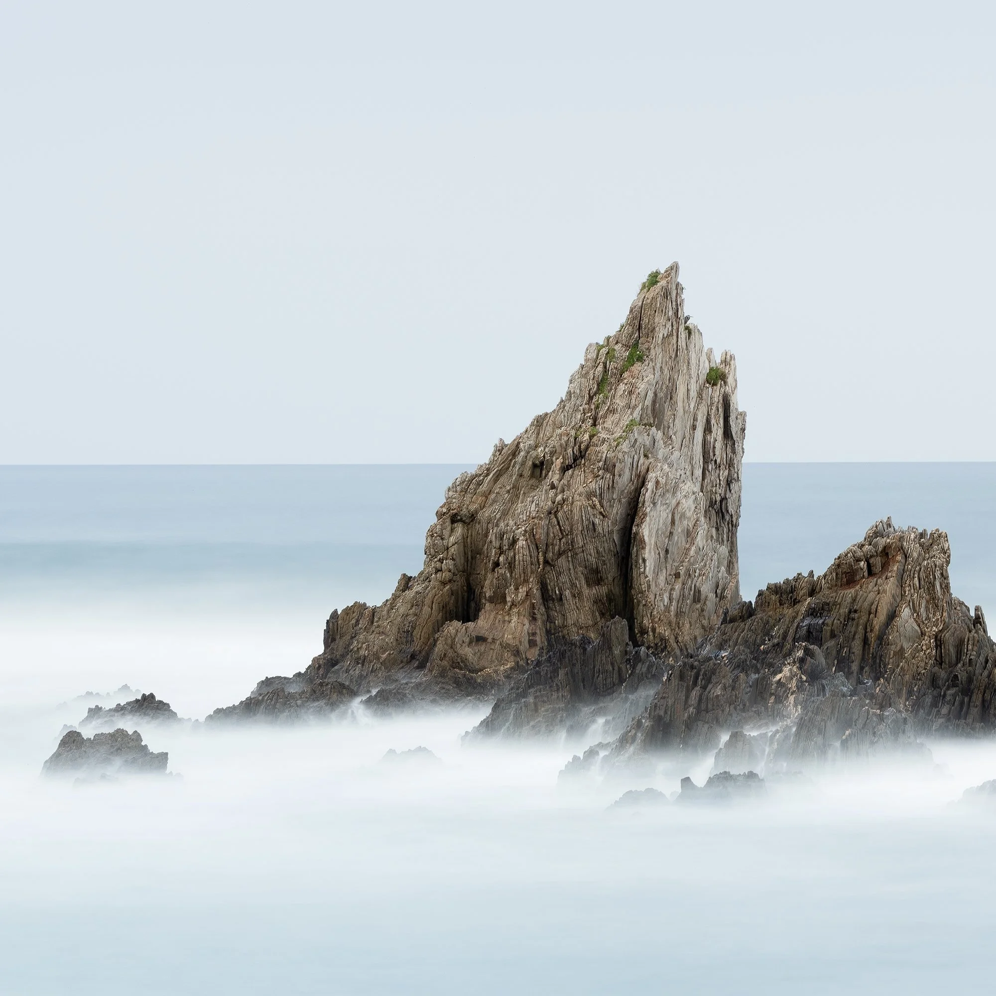 © 2023 Gerald Berghammer - Color seascape. Long exposure photograph of jagged sea stacks rising from the ocean with misty water and a light sky in the background. Print detail 1