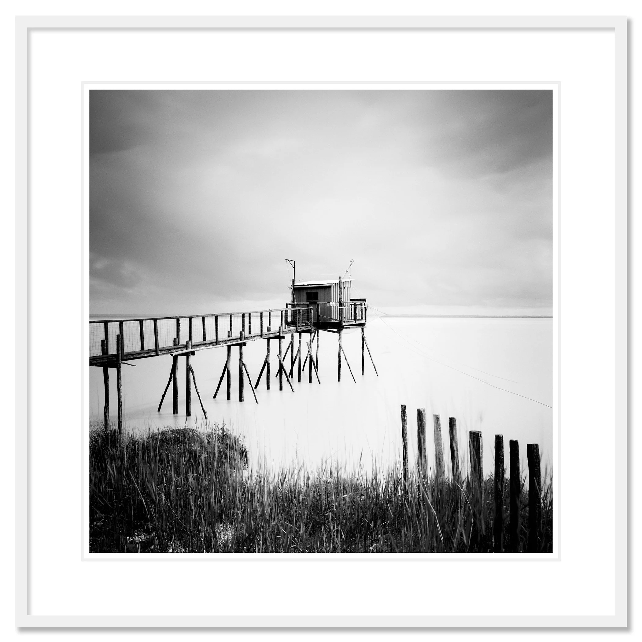 Gerald Berghammer - Black and white seascape photography. Small wooden hut on stilts connected by a long walkway over water with grass and a cloudy sky. Classci framed white