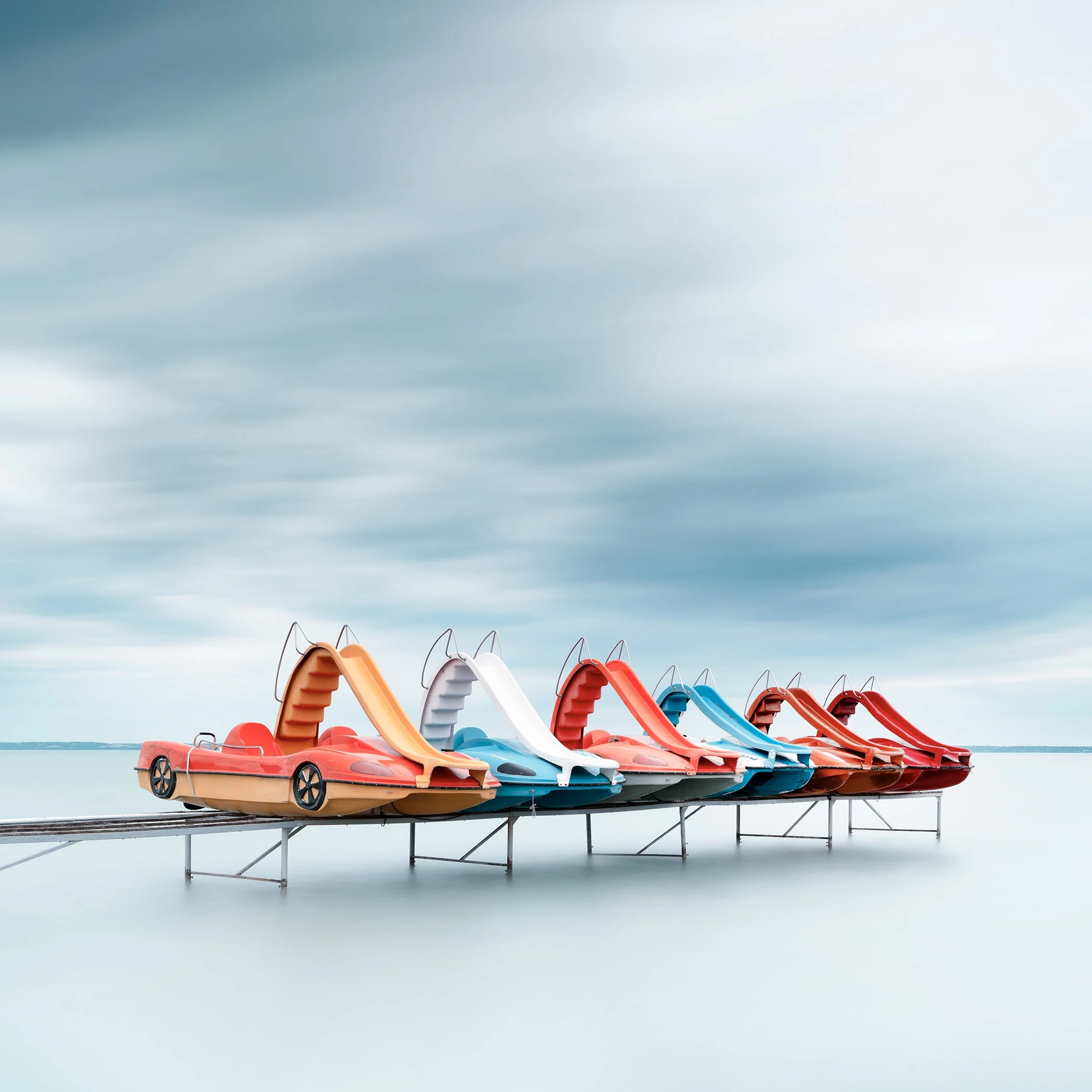 Gerald Berghammer - Color waterscape photography. A row of colorful plastic pedal boats shaped like race cars on a dock over water, under a cloudy sky.