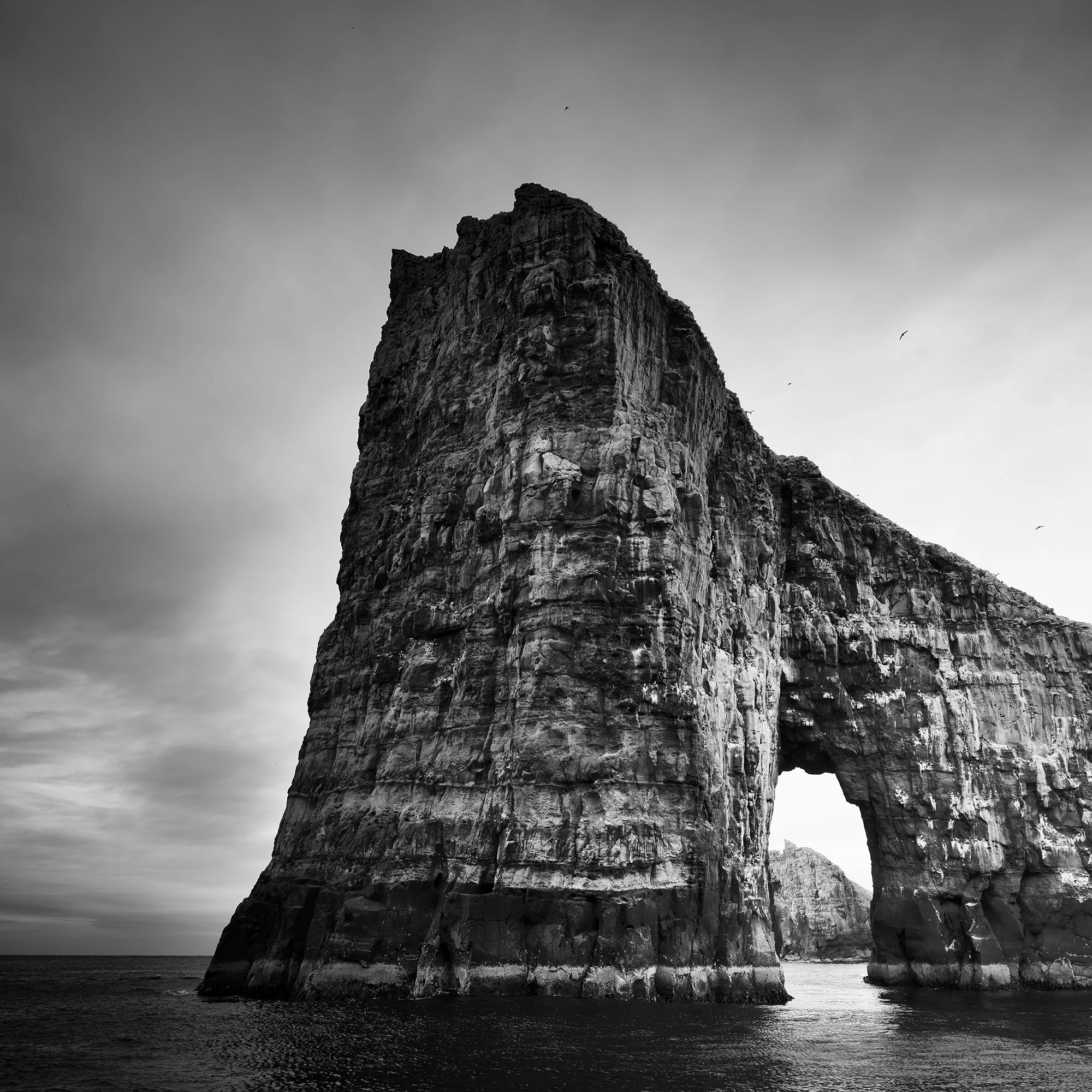 Gerald Berghammer - Black and white seascape photography.  Large natural rock formation with an arch in the middle, surrounded by water and a cloudy sky. Print detail 1